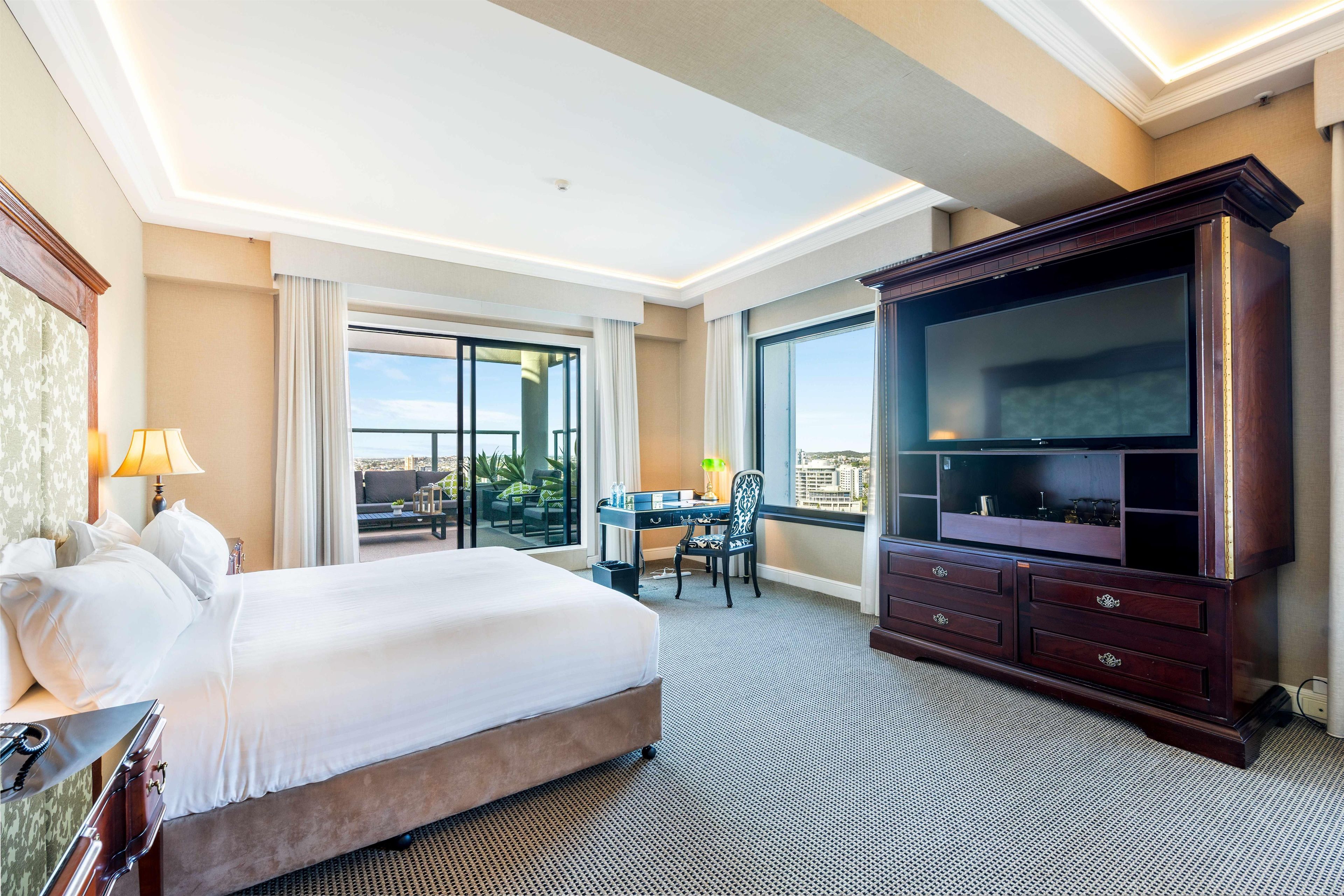 A hotel room with a large bed, a desk with a chair, a television, and a view of the city through the windows.