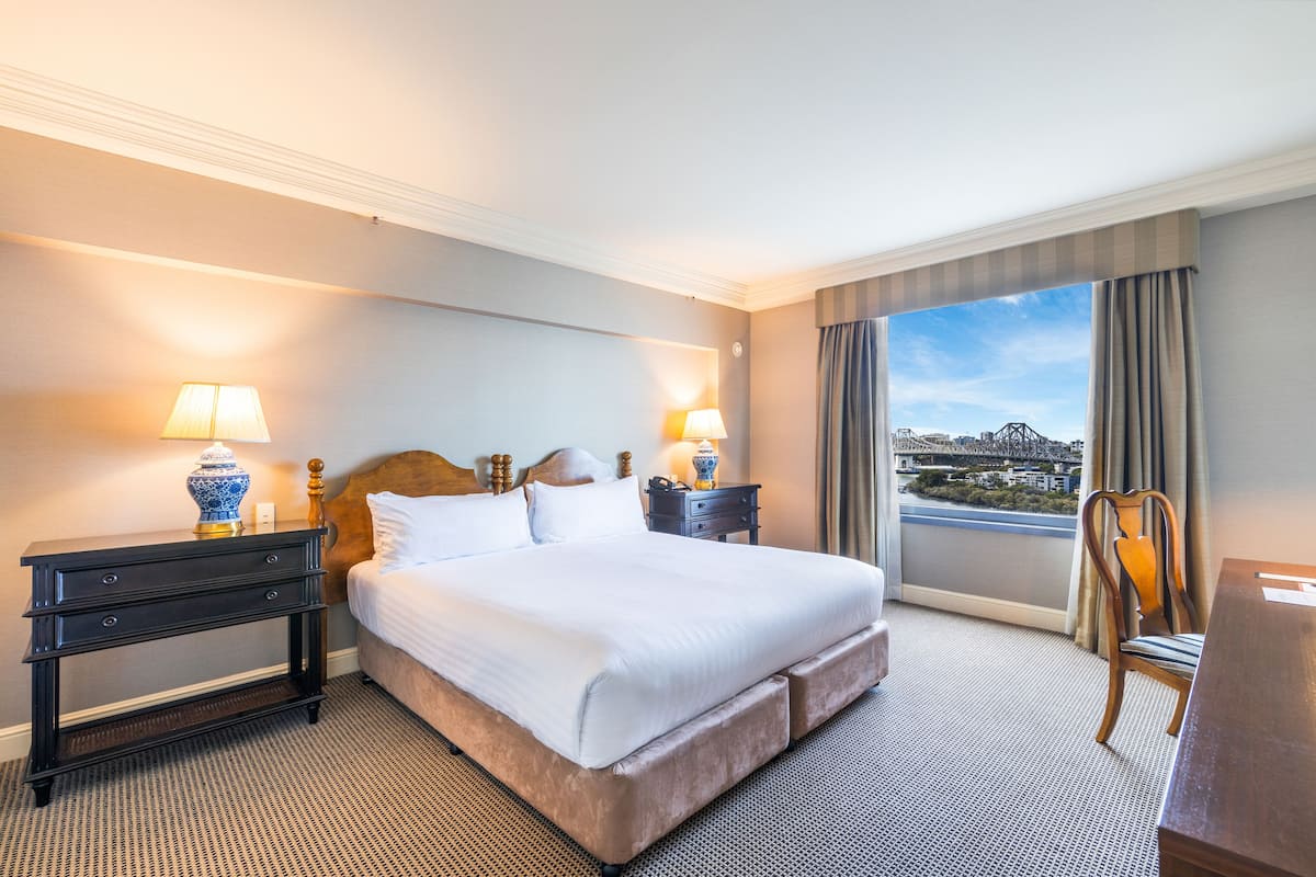 Een hotelkamer met een groot bed, twee nachtkastjes met lampen, een houten stoel en een uitzicht op een stadsbeeld door het raam.