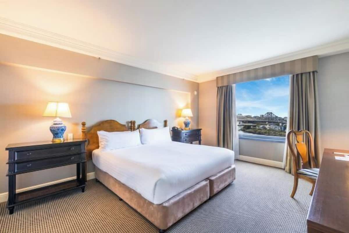 Een hotelkamer met een groot bed, twee nachtkastjes met lampen, een houten hoofdbord, uitzicht op een stadsbeeld door een groot raam, en een kleine tafel met een stoel.