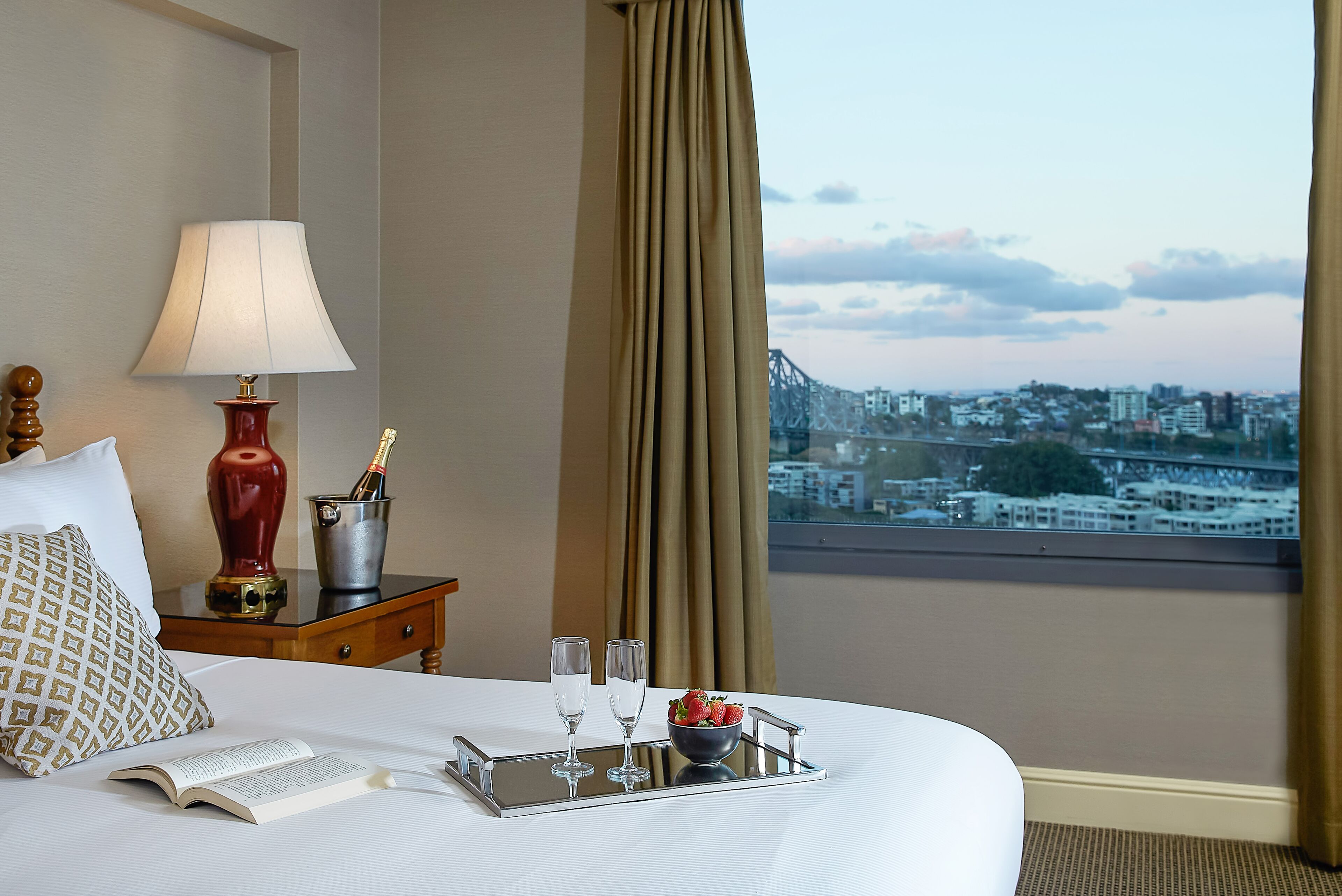 Habitación de hotel con cama, mesita de noche con lámpara, bandeja con vasos y fresas, y vista a la ciudad a través de la ventana.