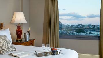 Habitación de hotel con cama, mesita de noche con lámpara, bandeja con vasos y fresas, y vista a la ciudad a través de la ventana.