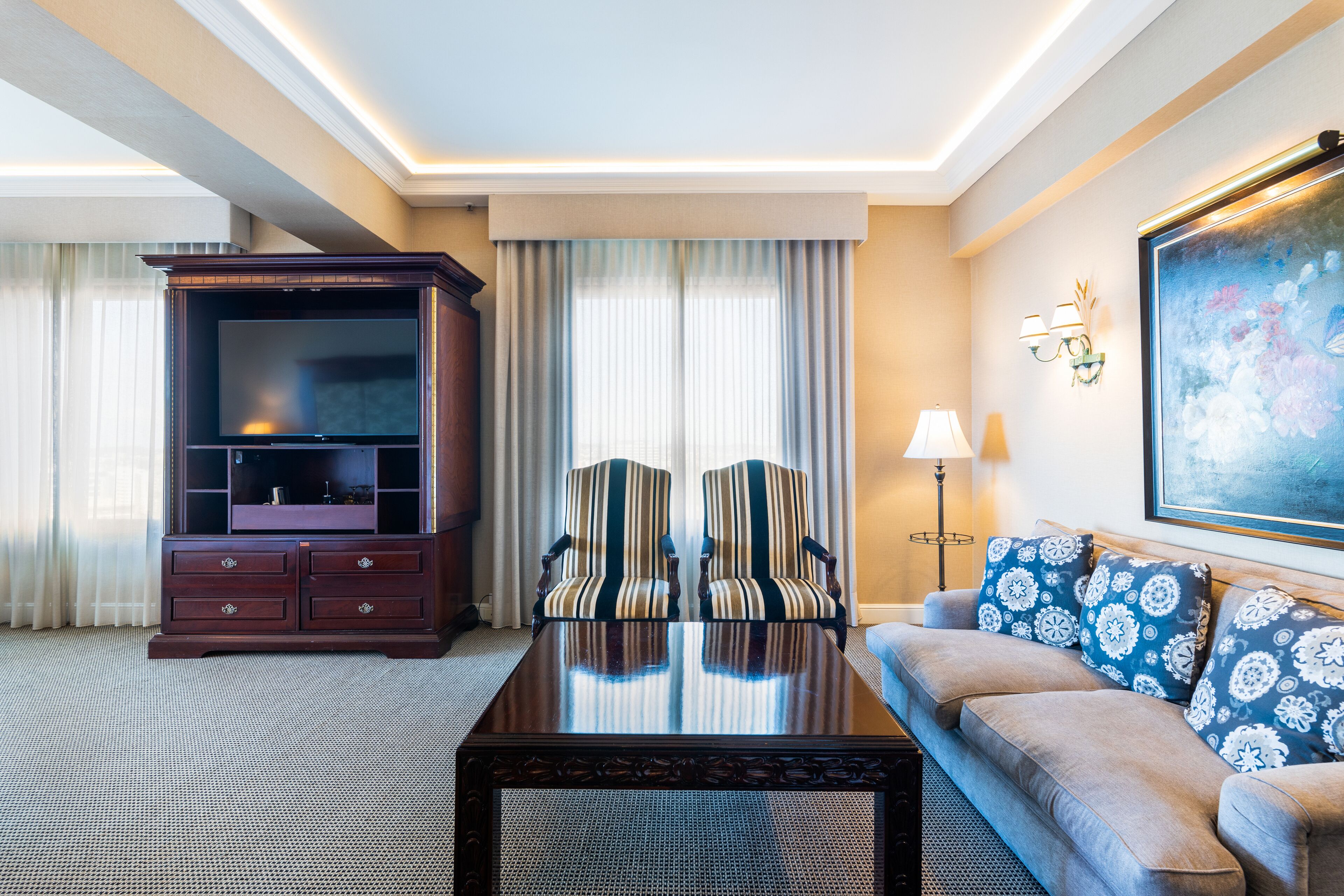 A hotel room with a large flat-screen TV, a sofa with patterned cushions, a wooden coffee table, and a painting on the wall.