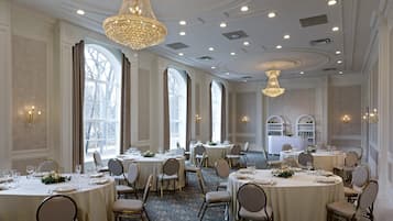 A formal dining room with multiple round tables, elegant chandeliers, and large windows.