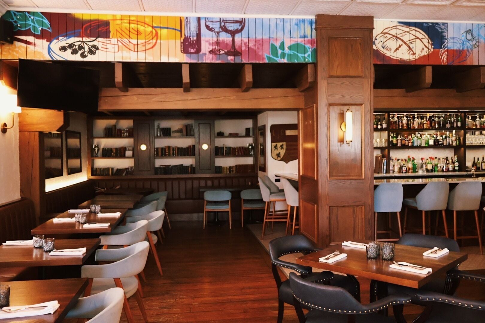A restaurant with wooden tables and chairs, a bar area with shelves of bottles, and a mural on the ceiling.
