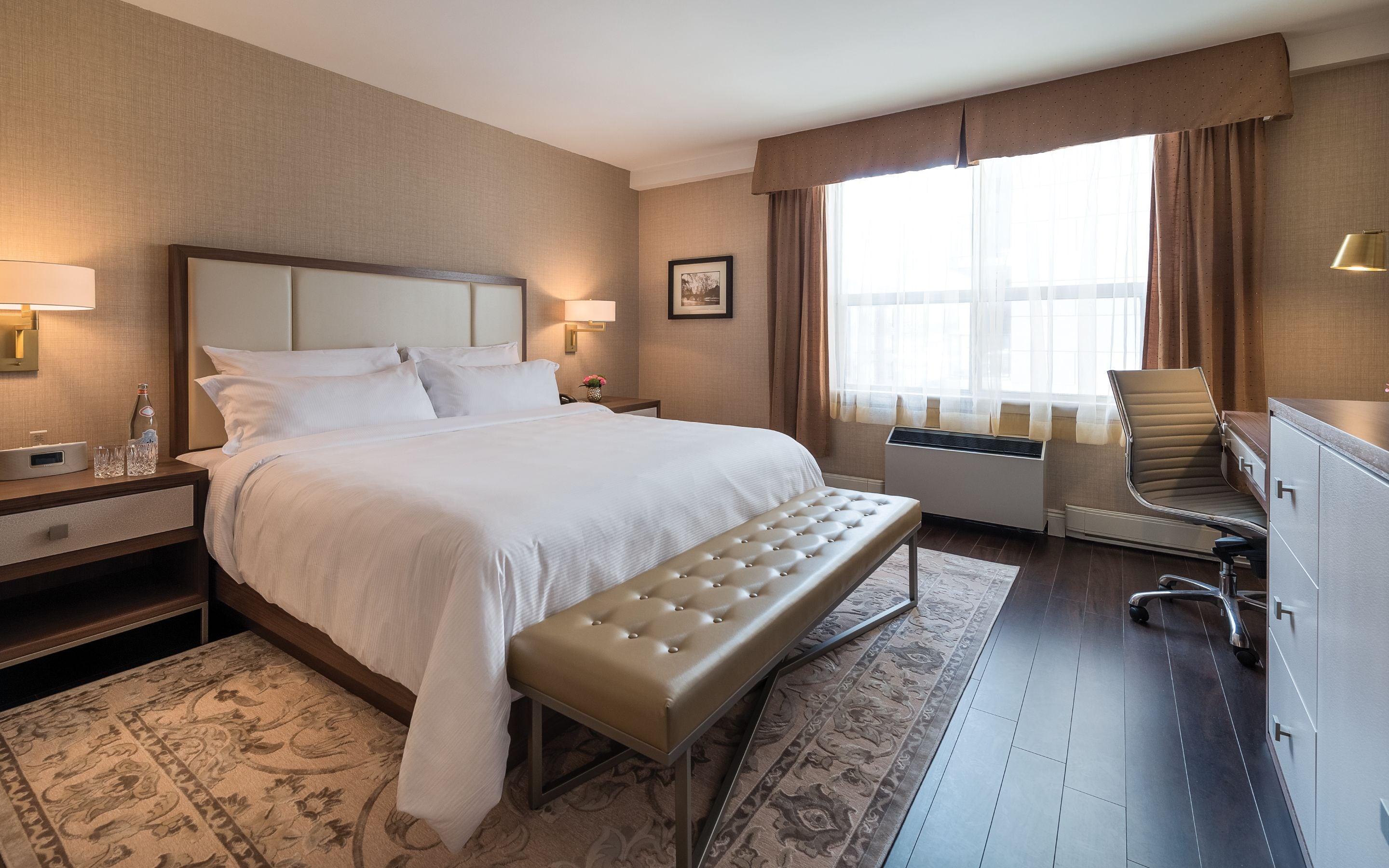 Habitación de hotel con una cama grande, un banco, un escritorio, una silla y una ventana con cortinas.