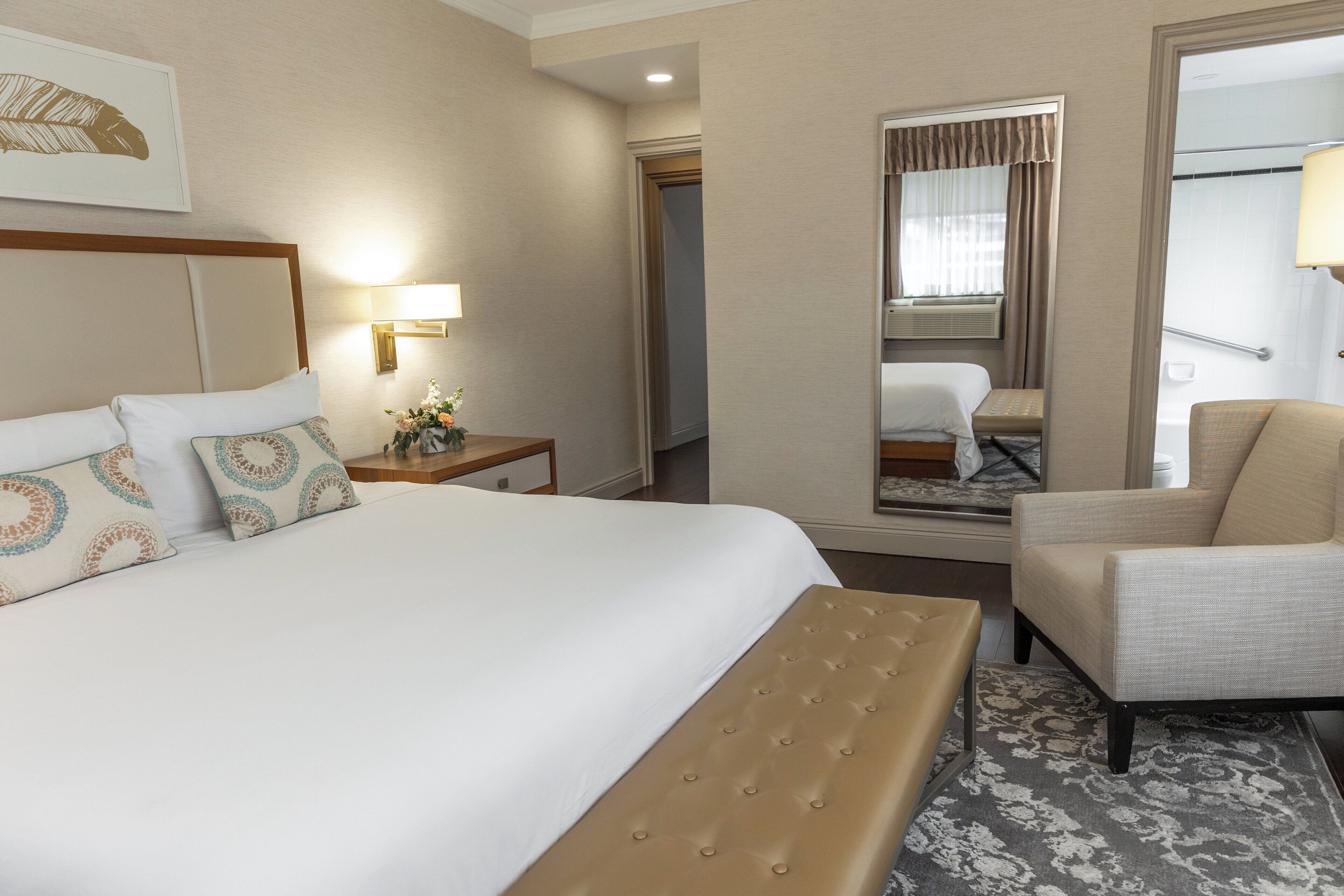 Una habitación de hotel con una cama grande, una zona de estar con una silla, una mesita con lámpara y una ventana con cortinas.