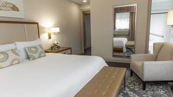 Una habitación de hotel con una cama grande, una zona de estar con una silla, una mesita con lámpara y una ventana con cortinas.