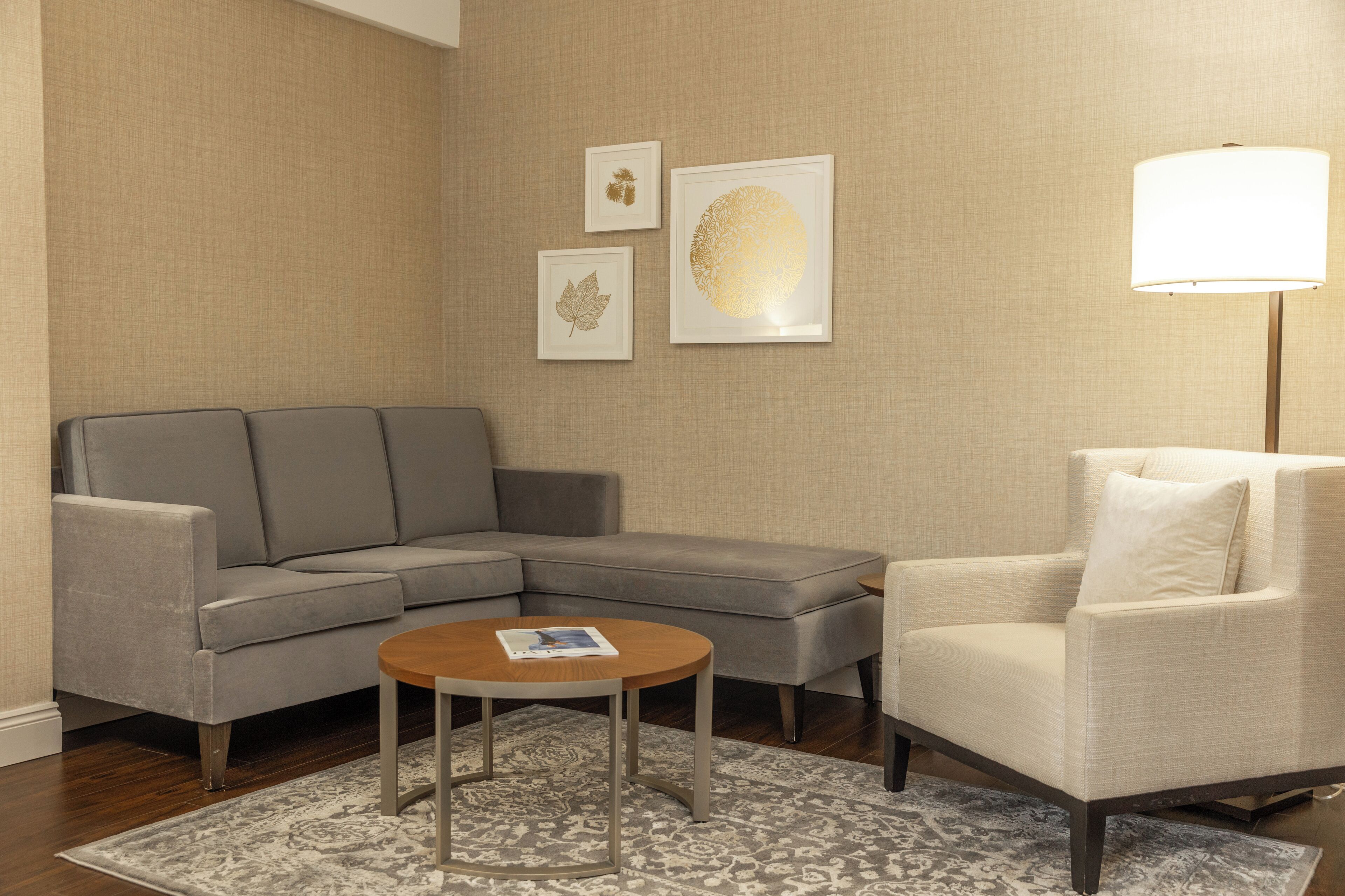 A living room with a gray sectional sofa, a round coffee table, a white armchair, and a floor lamp. There are three framed artworks on the wall.