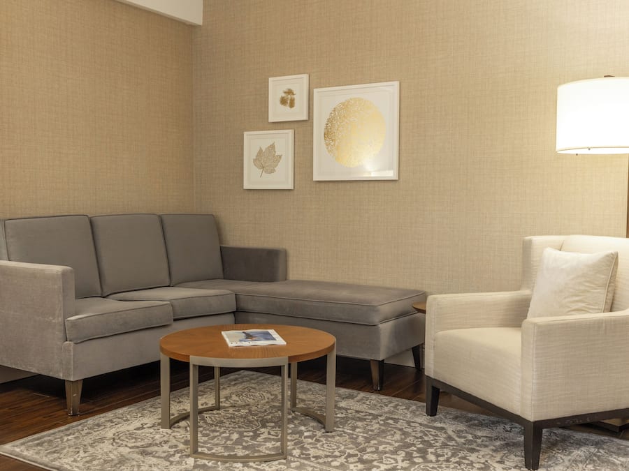 A living room with a gray sectional sofa, a round coffee table, a white armchair, and a floor lamp. There are three framed artworks on the wall.