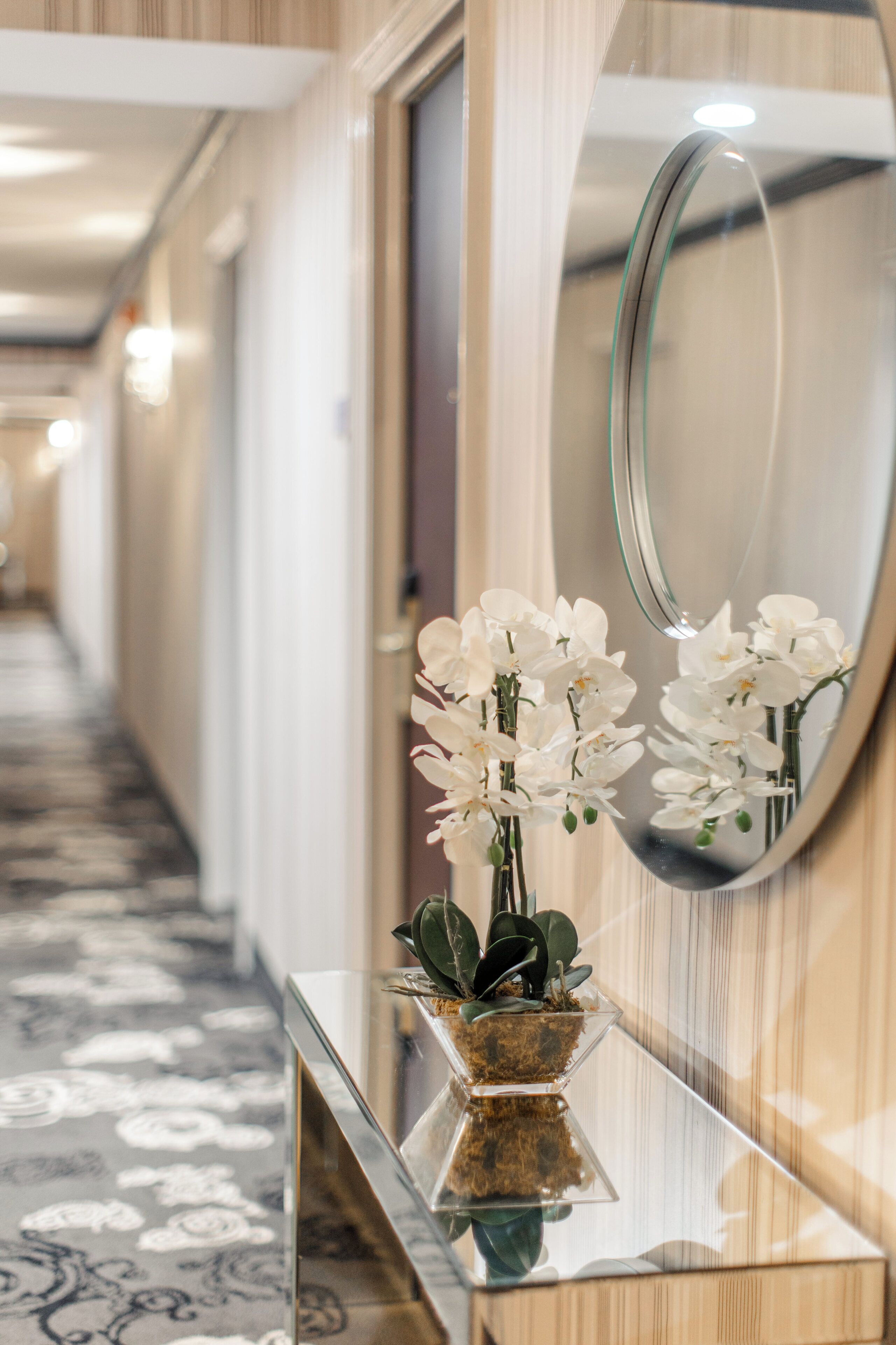 A hallway with a mirror reflecting white flowers and a glass vase on a wooden counter.