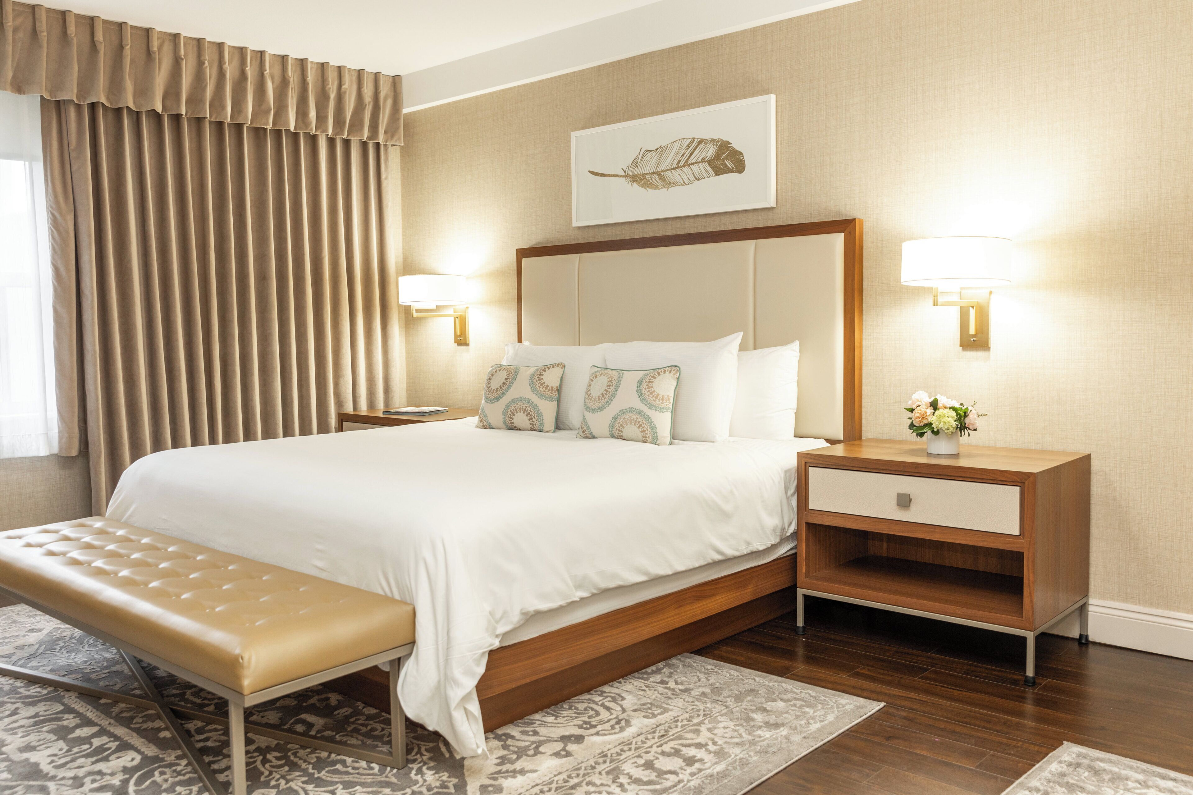 A neatly arranged hotel room with a large bed, a bench, a nightstand with a lamp, and a wall-mounted artwork.