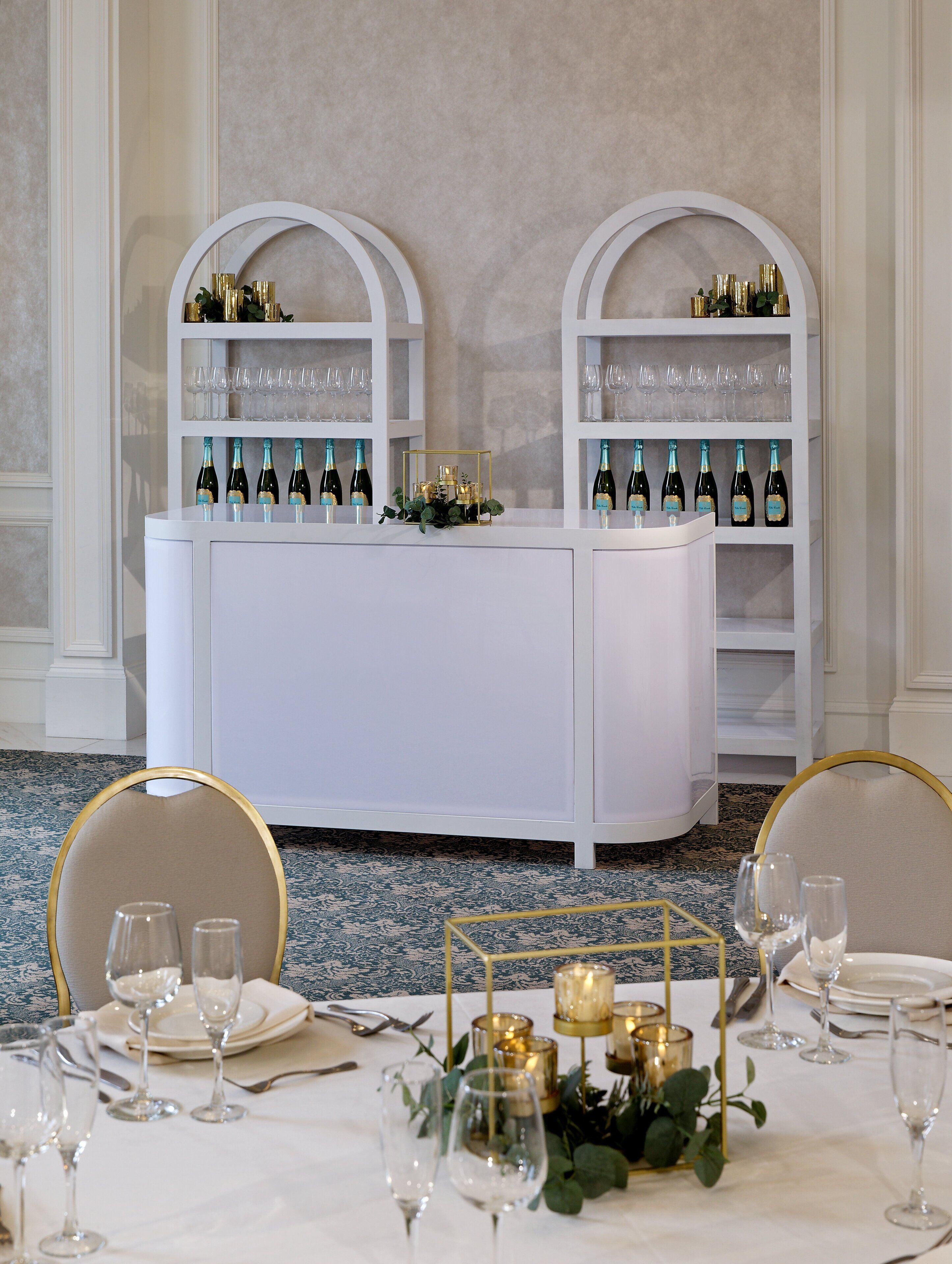 A formal dining setup with a white table, gold-accented chairs, and a white bar area with wine bottles and glasses.