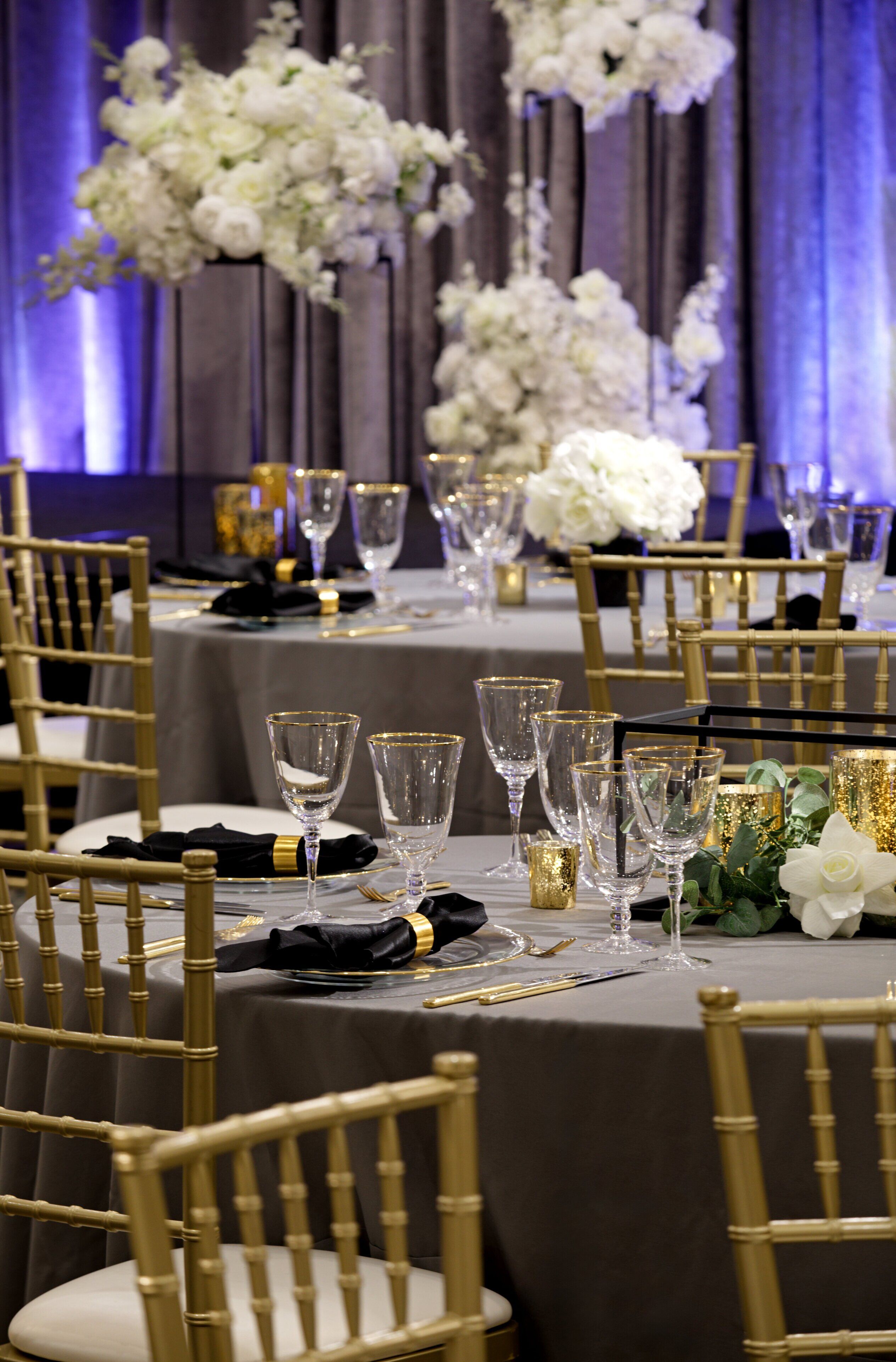 Una mesa de banquete dispuesta con sillas doradas, flores blancas y cristalería transparente.