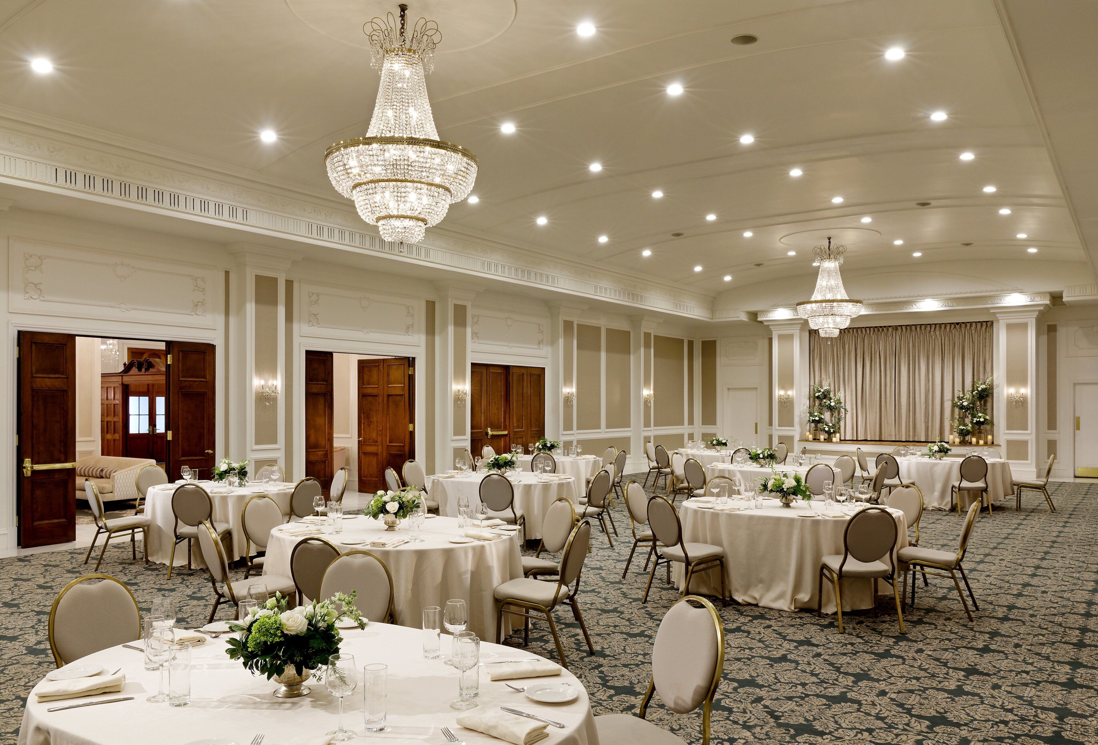 A banquet hall with round tables, chairs, chandeliers, and a carpeted floor.