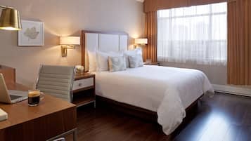 A hotel room with a large bed, two bedside tables with lamps, a desk with a laptop and a coffee cup, a chair, and a window with curtains.