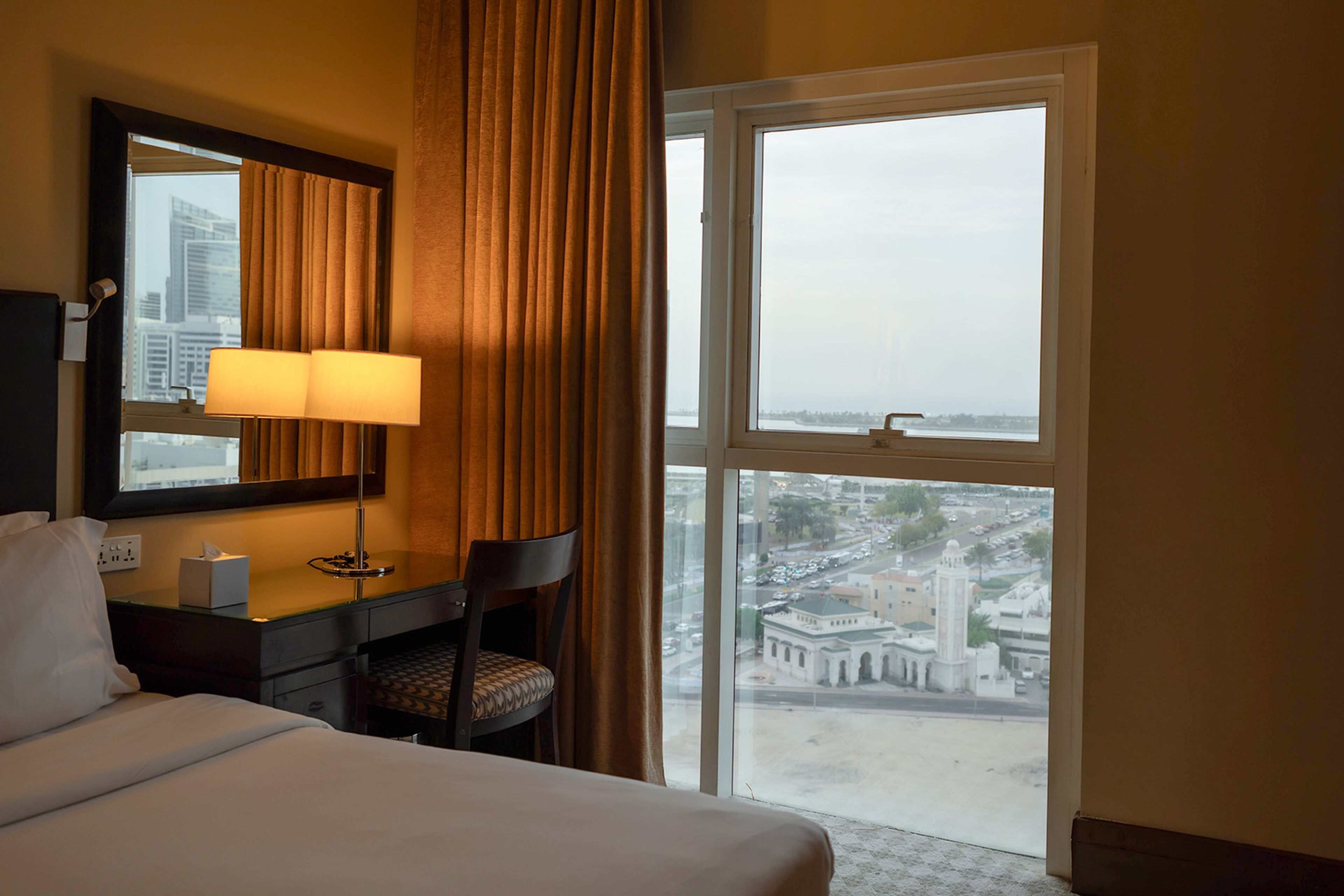 Una habitación de hotel con cama, escritorio con lámpara, silla y un amplio ventanal con vista a un paisaje urbano y un edificio.