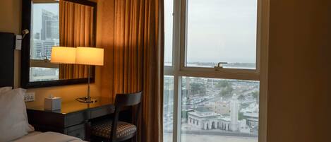 Una habitación de hotel con cama, escritorio con lámpara, silla y un amplio ventanal con vista a un paisaje urbano y un edificio.
