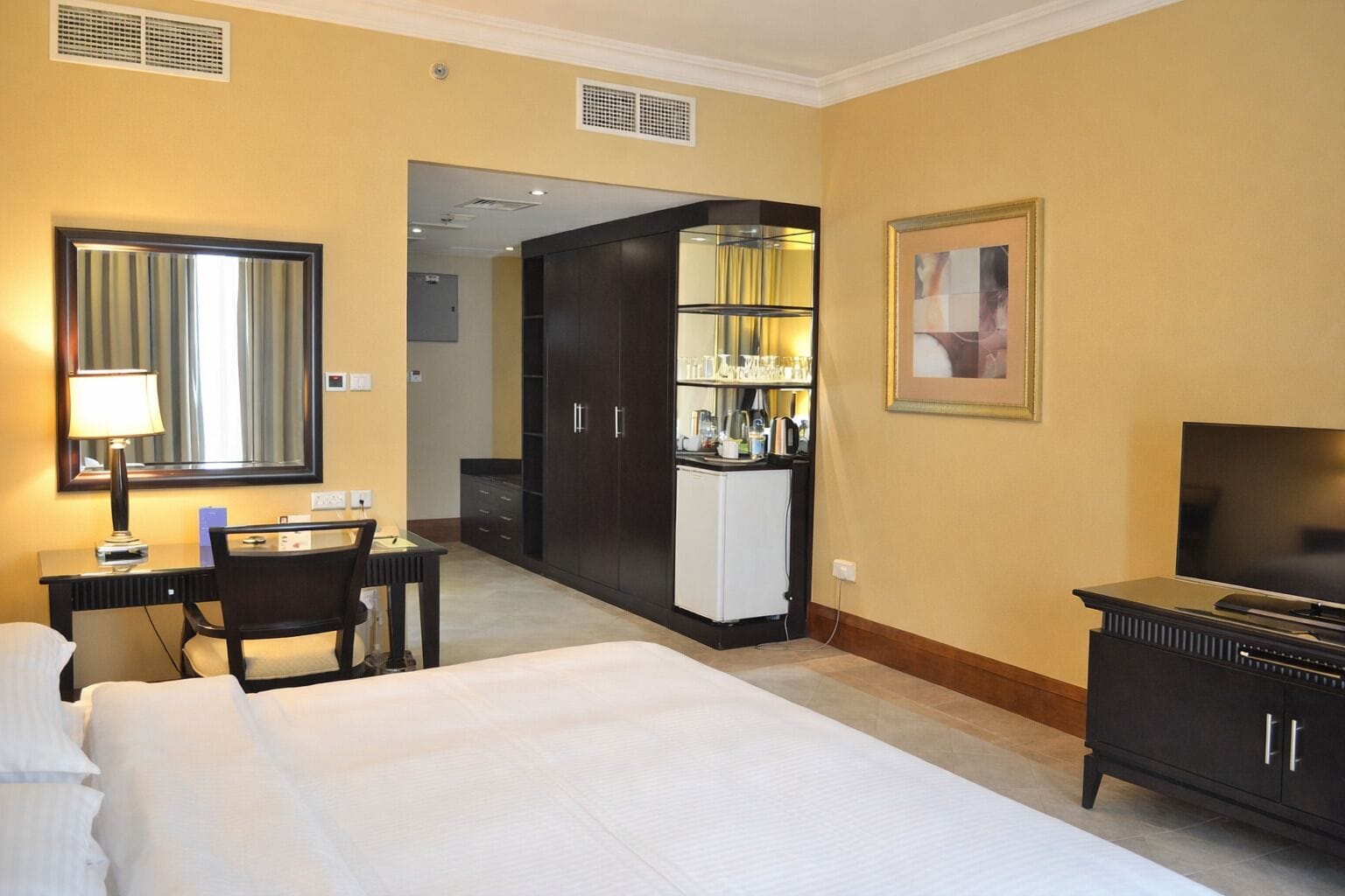 Habitación de hotel con cama, escritorio con silla, televisor, minibar y un espejo grande.