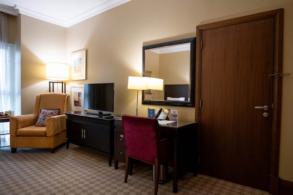 A hotel room with a desk, two chairs, a television, a telephone, a mirror, and a door.