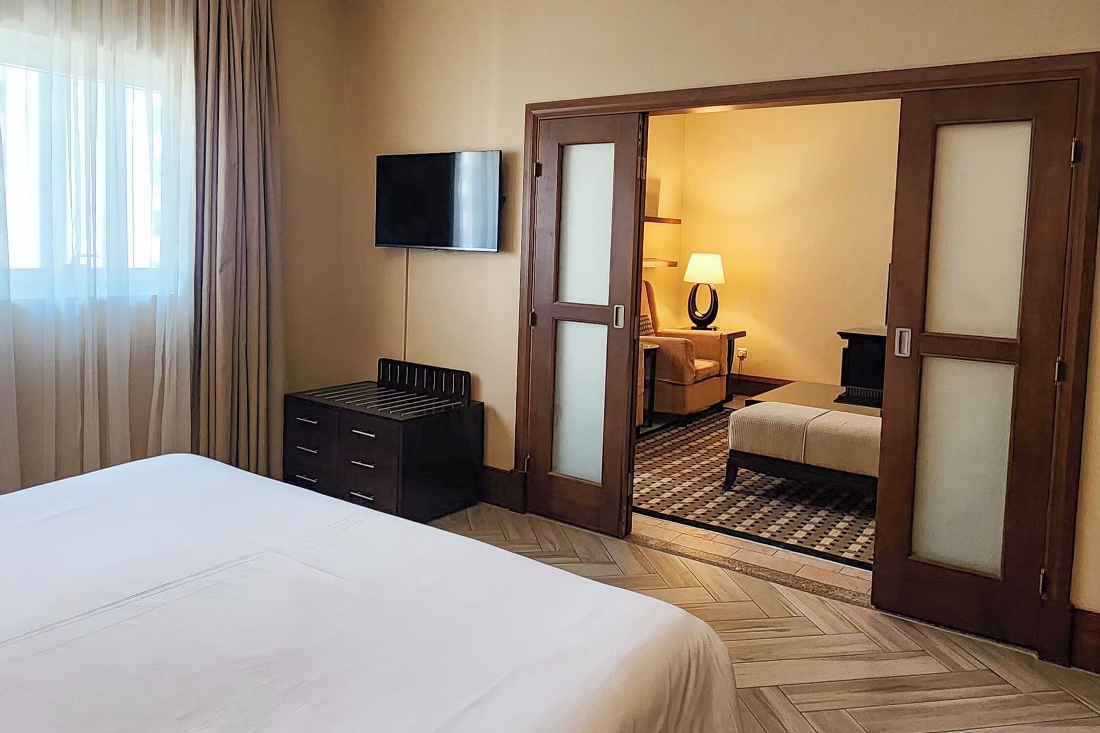 A hotel room with a bed, a TV, a desk with a lamp, and a door leading to another room.
