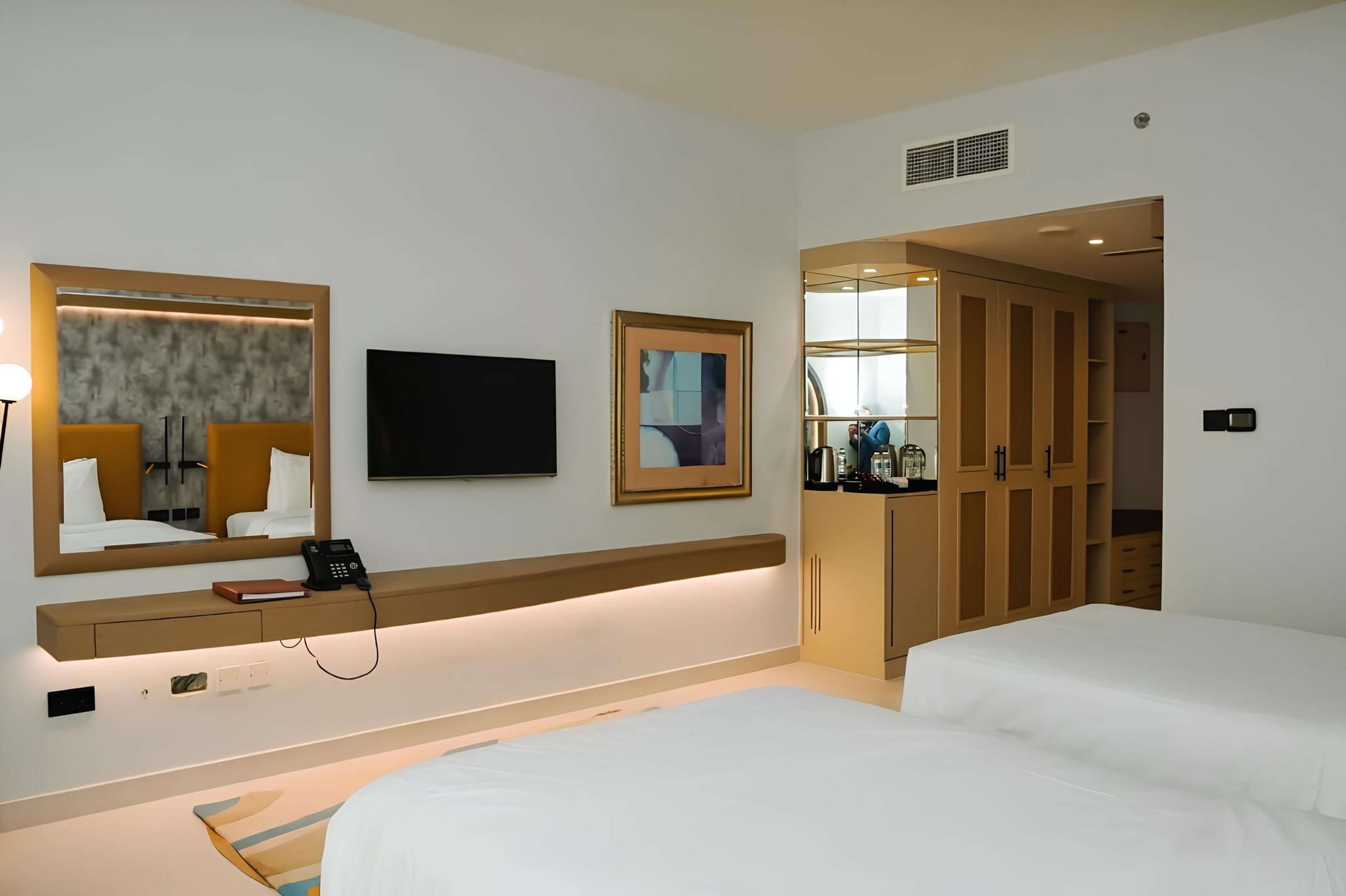A hotel room with a bed, a bedside table with a lamp, a phone, a TV, a framed picture, and a wardrobe.