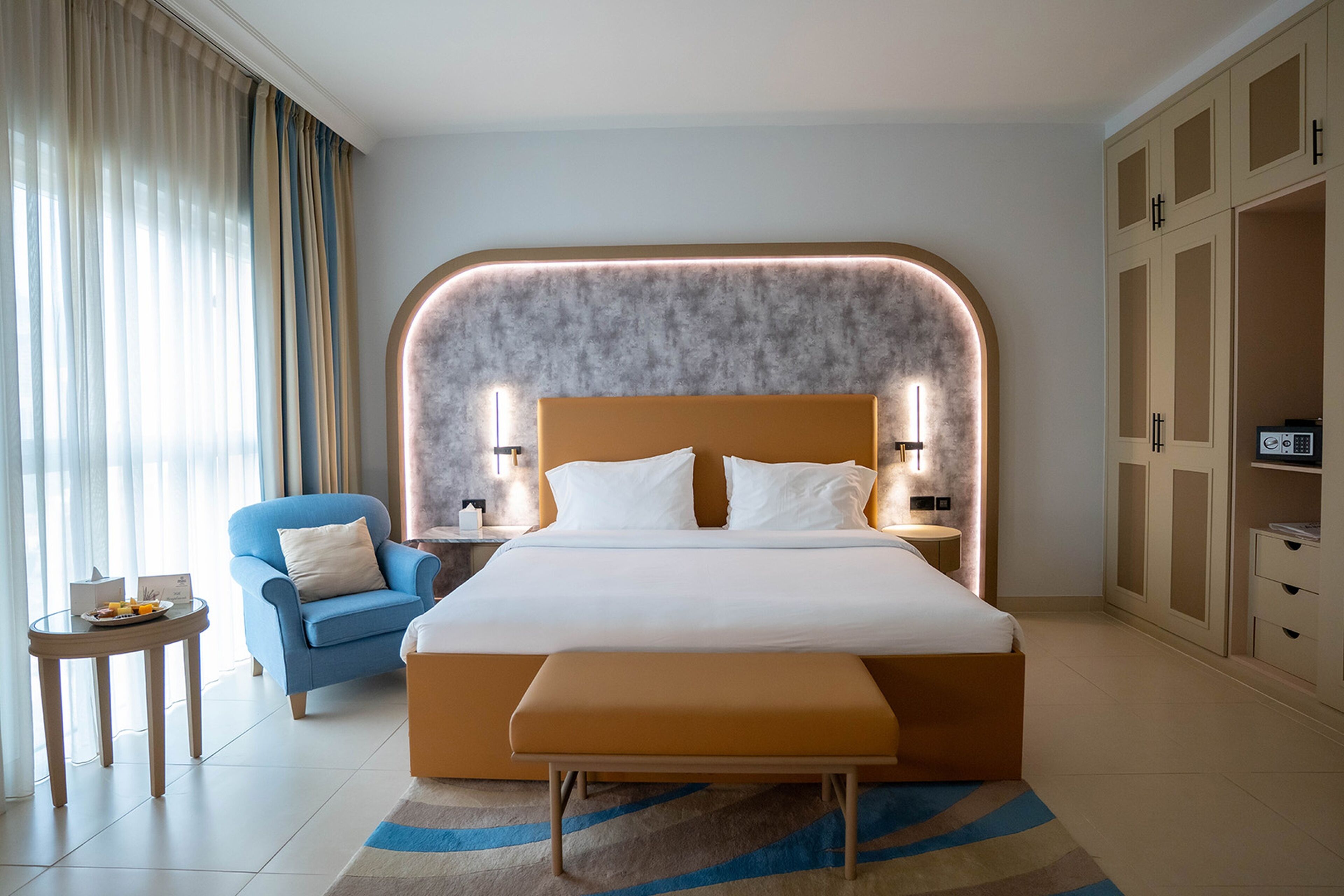 A modern hotel room with a large bed, a blue armchair, a small table with a fruit plate, and a wardrobe.