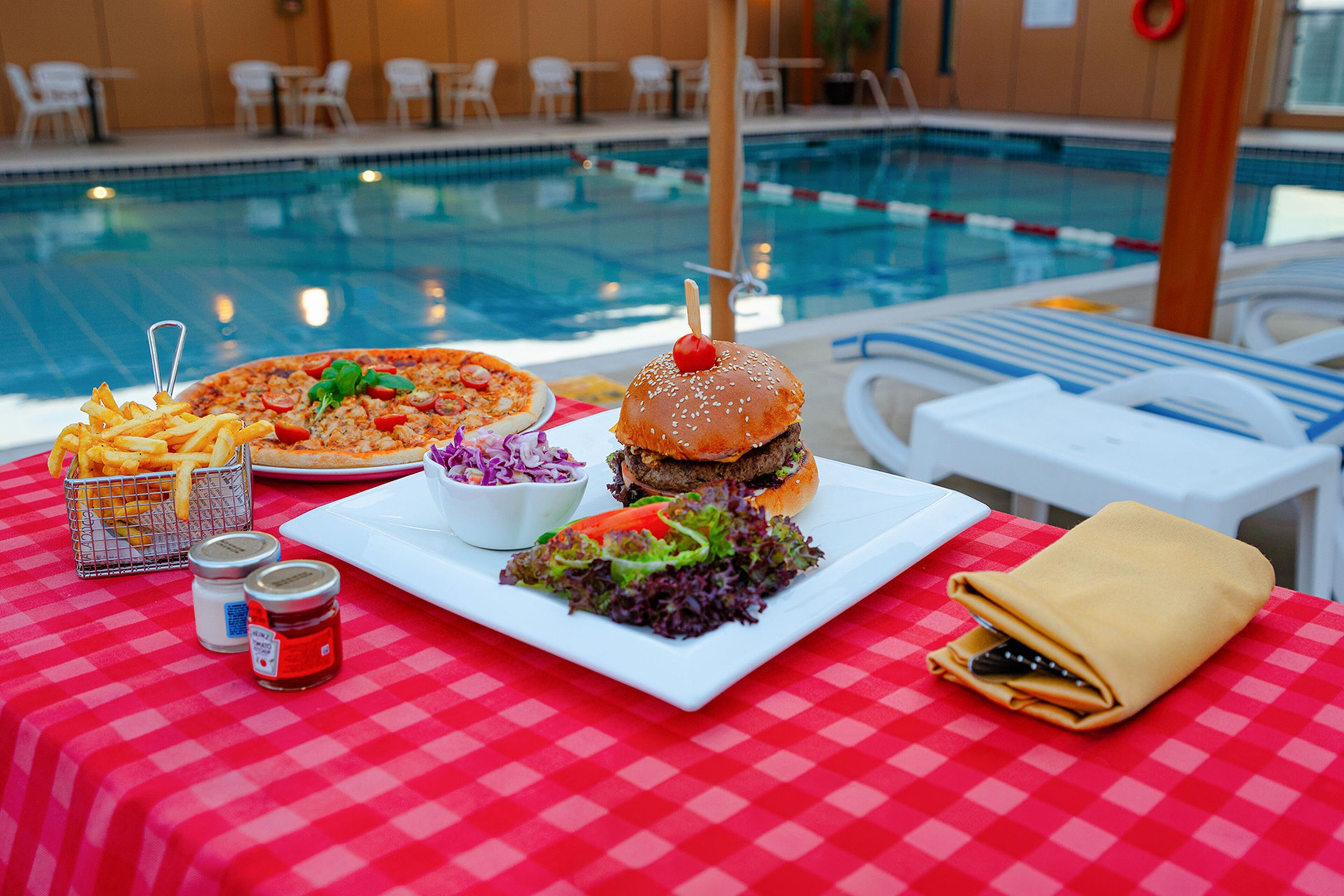 Une table à manger recouverte d’une nappe à carreaux, avec une pizza, un hamburger, des frites, une salade et divers condiments.