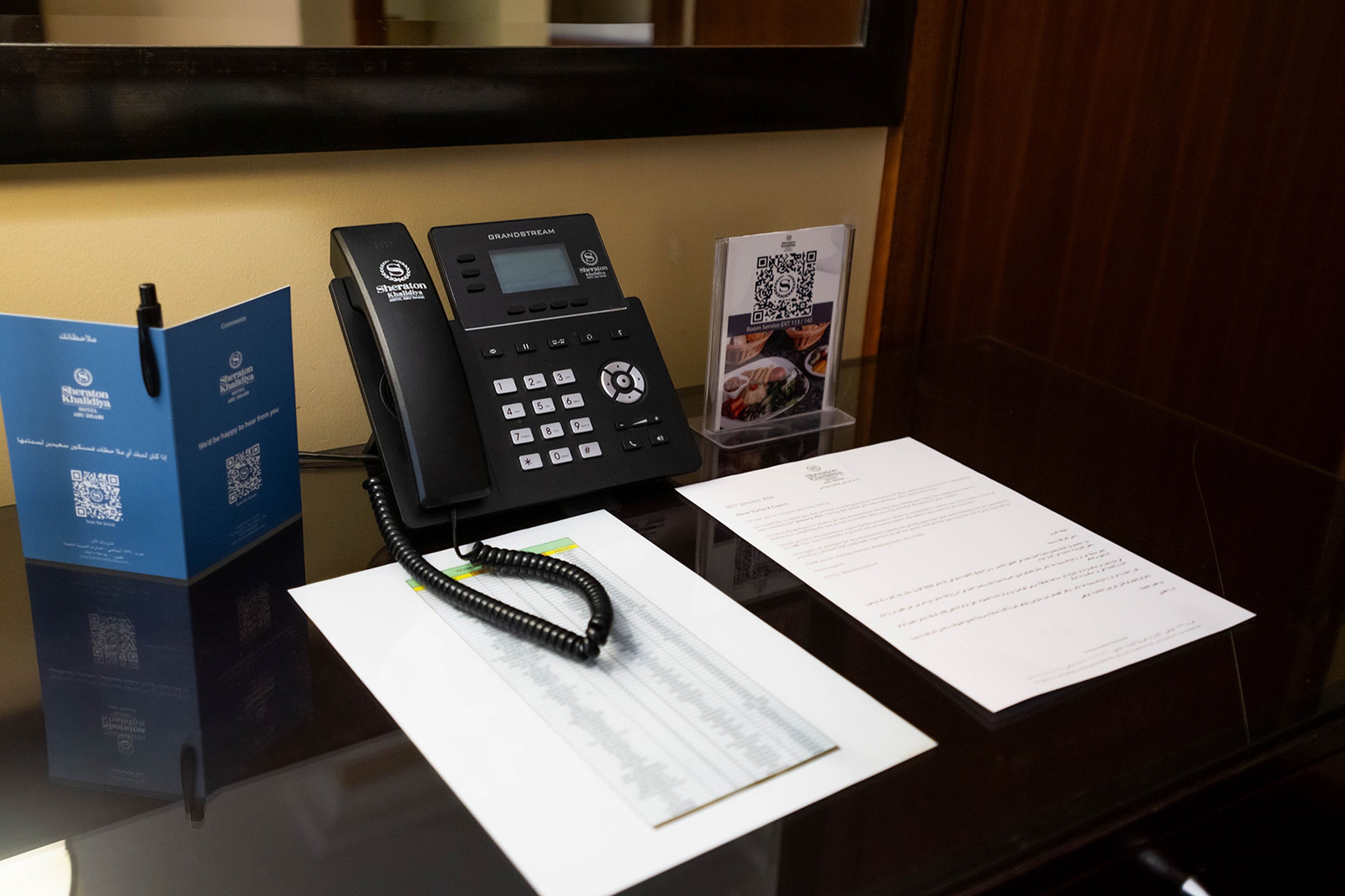 Zona de recepción de hotel con un teléfono negro, un folleto azul, un soporte para código QR y algunos documentos sobre la mesa.
