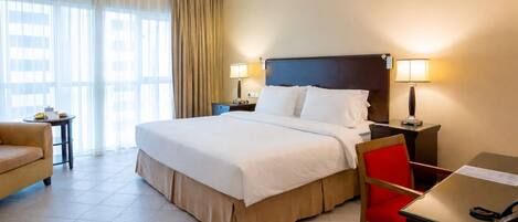 Une chambre d’hôtel avec un lit, des tables de chevet, un bureau, une chaise et une fenêtre avec des rideaux.