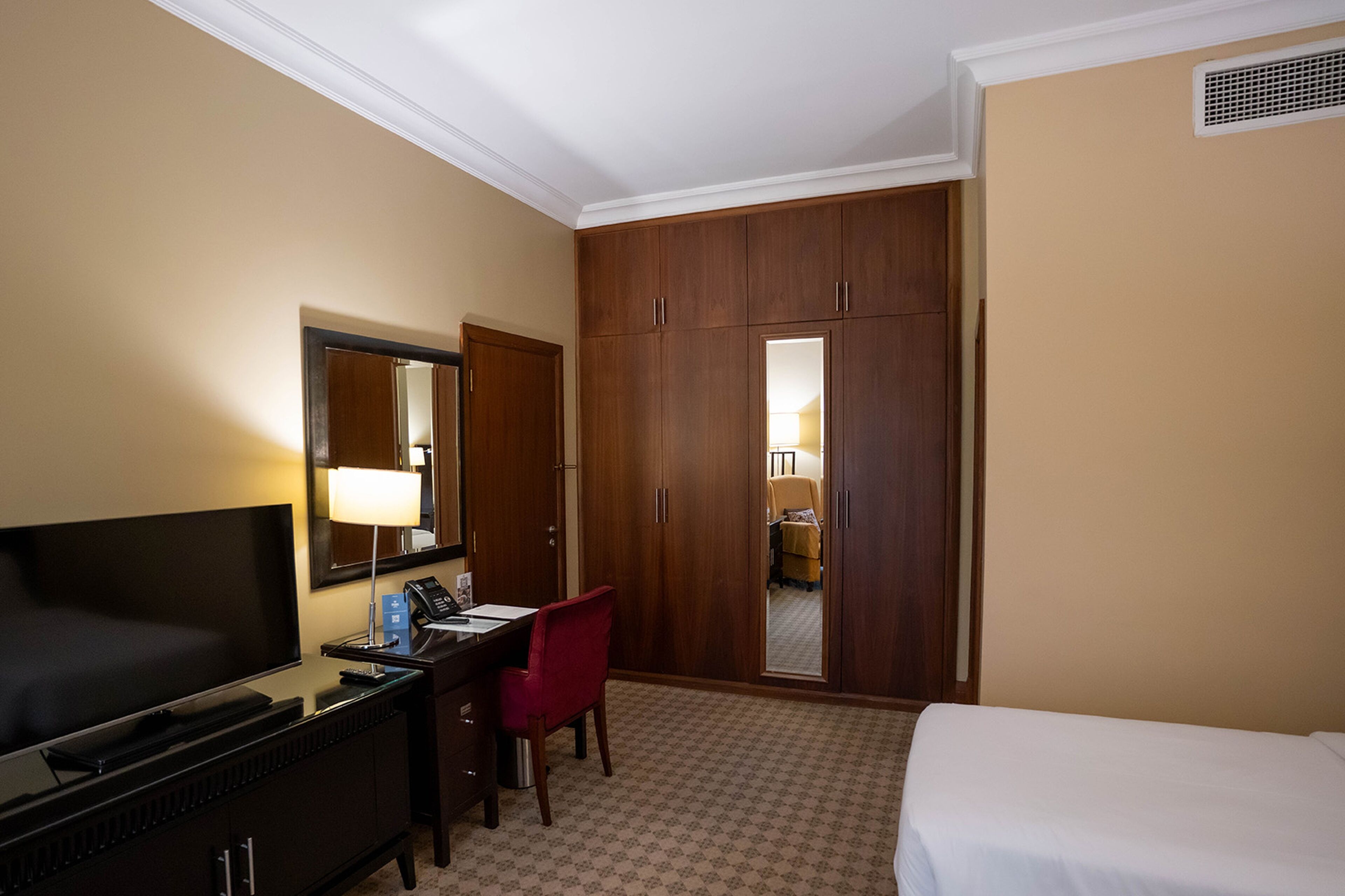 Habitación de hotel con cama, escritorio, silla, televisor, armario y espejo.