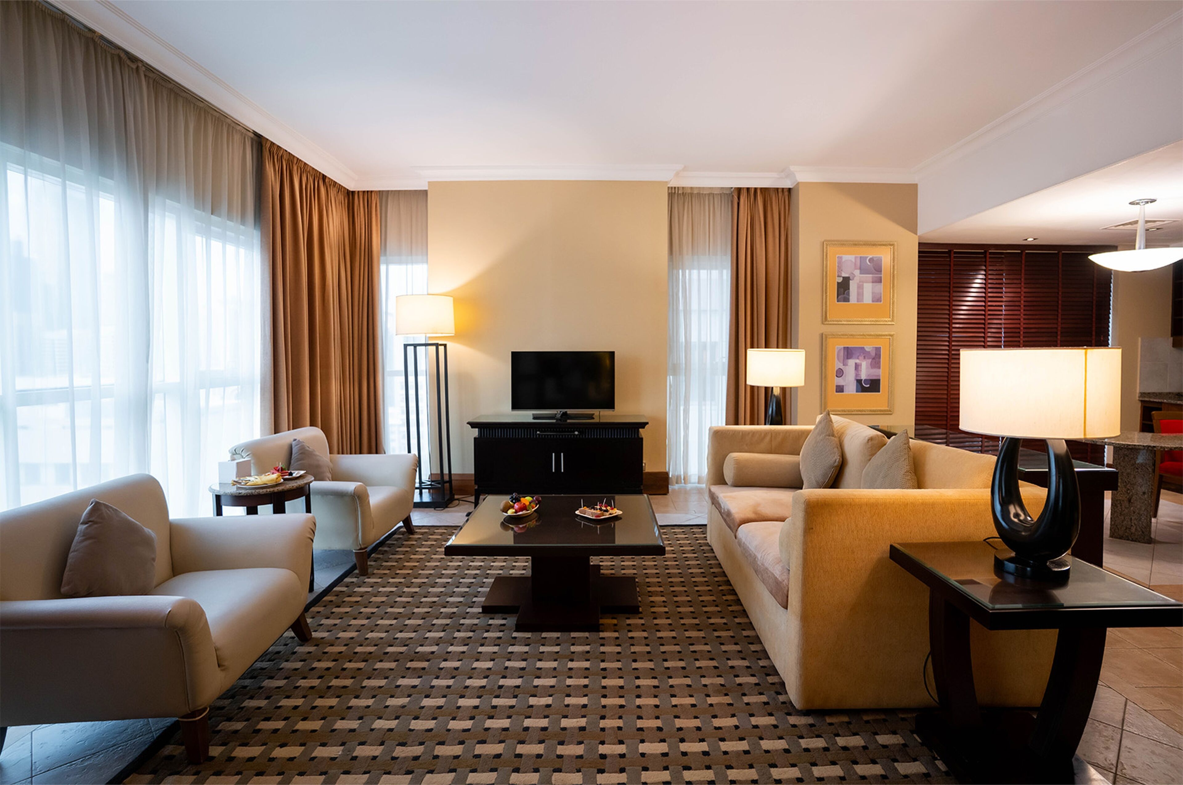 A hotel living room with a sofa, armchairs, a coffee table, a TV, and a lamp.