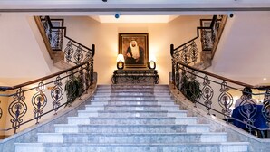 A grand staircase with marble steps and ornate metal railings, leading to a landing with a framed portrait and two wall-mounted lamps. - Sheraton Khalidiya Hotel (Abu Dhabi)
