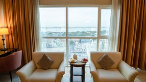A room with two beige sofas, a small table with a plate of food, and a view of a cityscape through large windows. - Sheraton Khalidiya Hotel (Abu Dhabi)