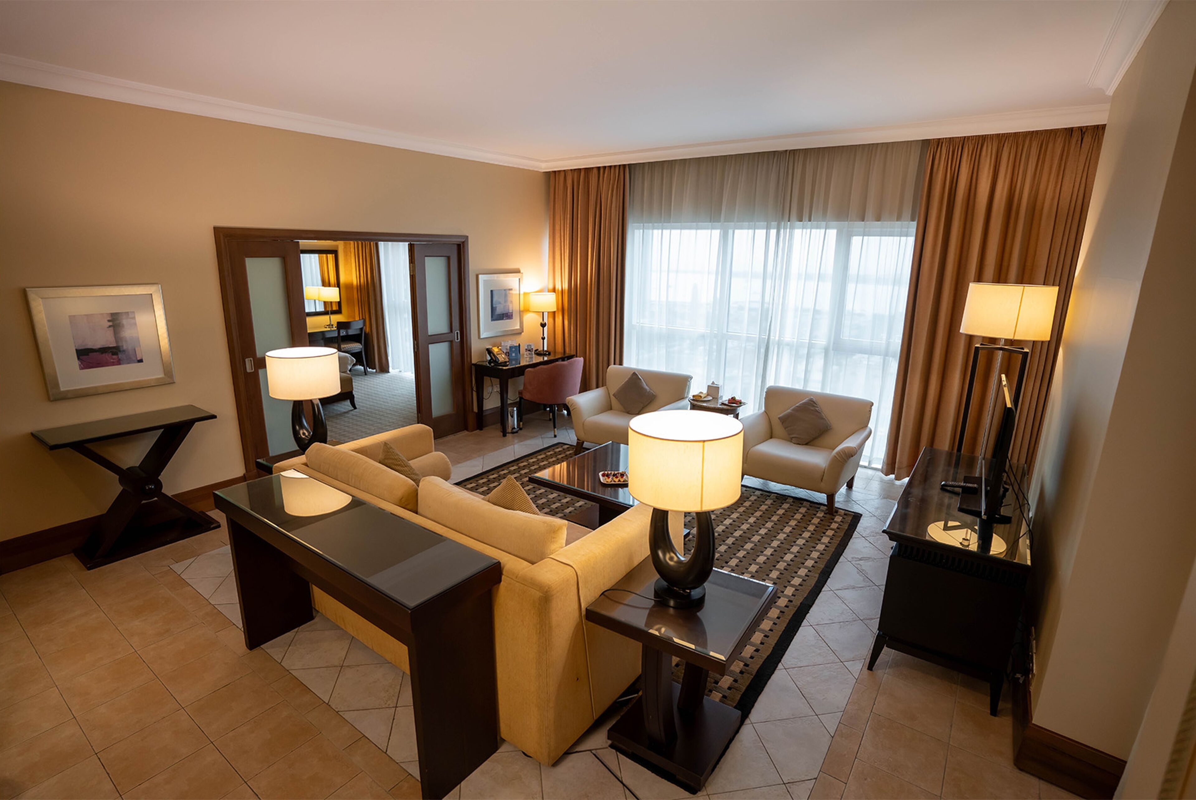 Hotelkamer met een bank, fauteuil, salontafel, lampen, een spiegel en uitzicht op de stad door grote ramen.