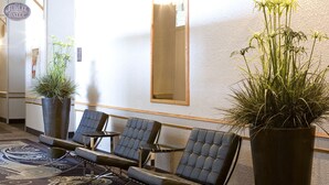 A hotel lobby with a patterned carpet, seating area, large potted plants, and a reception desk.