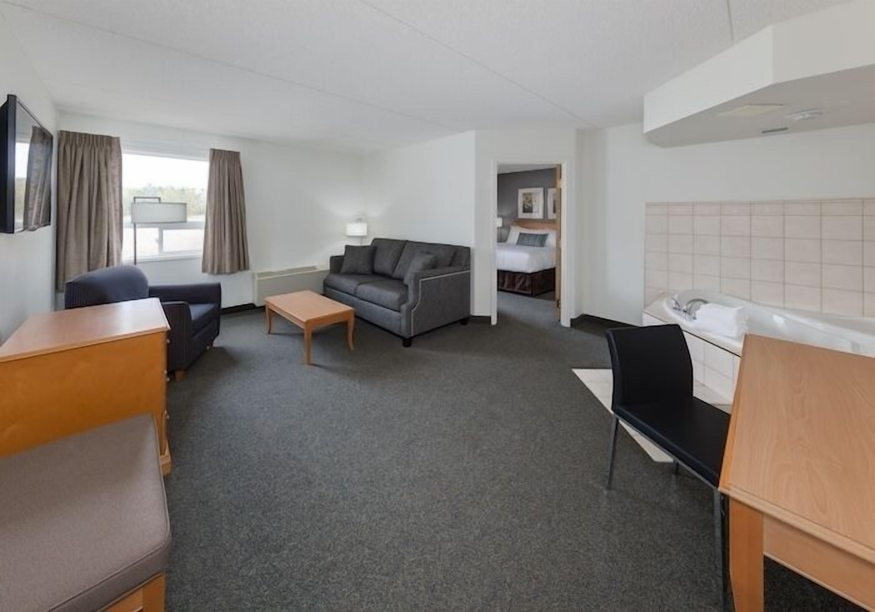 Habitación de hotel con alfombra gris, sofá gris, escritorio de madera, silla negra, banco de madera, mesita de noche, cama y ventana con cortinas, además de un cabecero blanco.