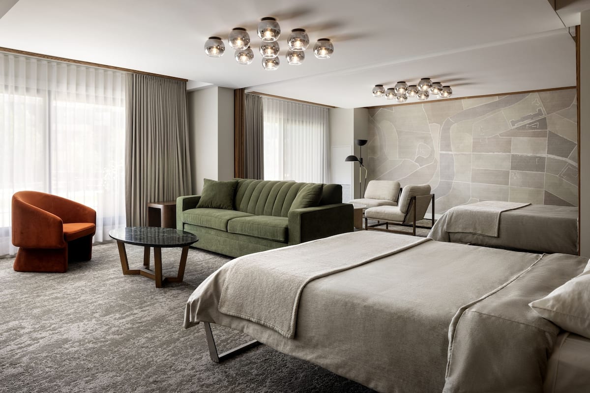 A modern hotel room with a large bed, a green sofa, an orange armchair, and a small round table.