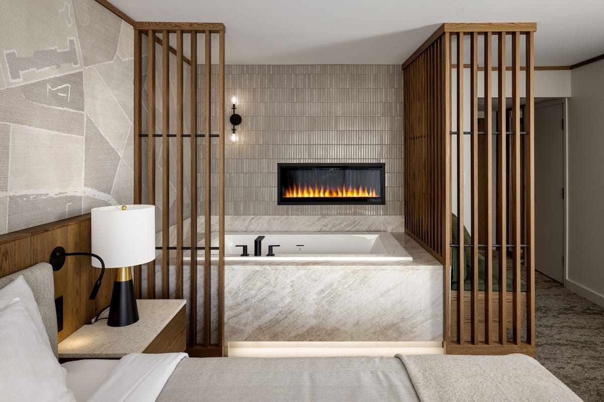 A modern hotel room with a fireplace, a large bed, a sitting area, and a bathroom with a bathtub.