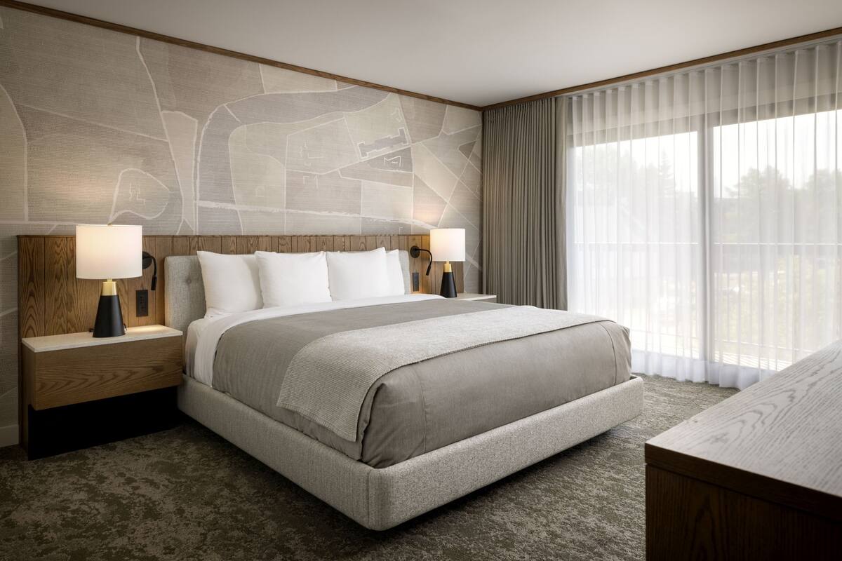 A hotel room with a large bed, two bedside tables with lamps, a headboard with a map design, and a large window with curtains.