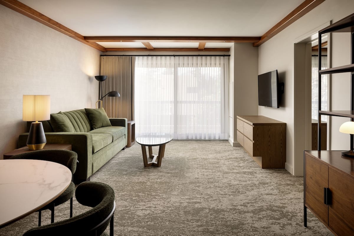 A modern hotel room with a green sofa, a round table, a small coffee table, a TV mounted on the wall, and a wooden cabinet.