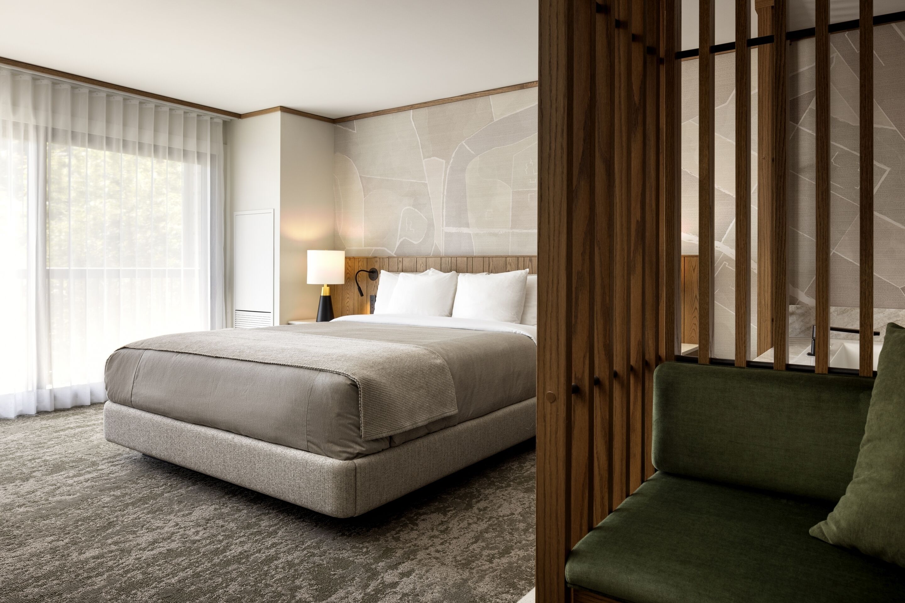 Una habitación de hotel con una cama grande, un sofá verde, un cabecero de madera y un ventanal con cortinas traslúcidas.