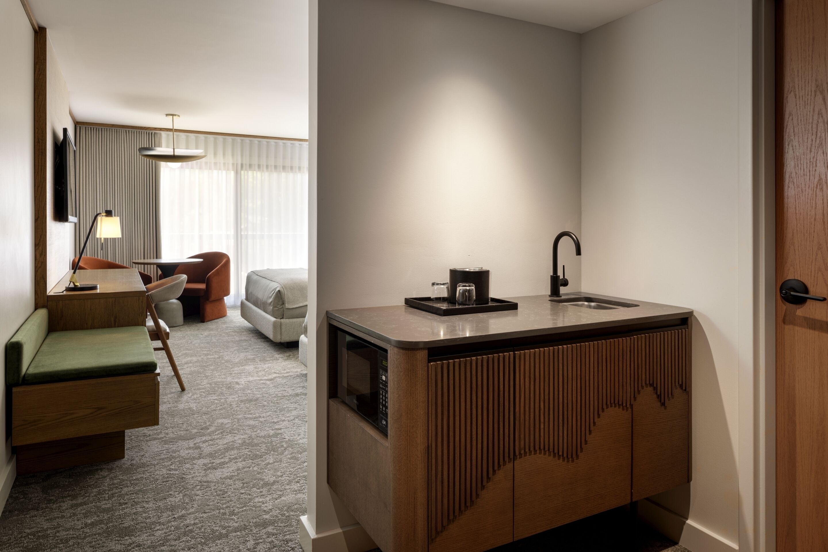 Habitación de hotel compacta con kitchenette, zona de estar con sofá y sillones, y un ventanal grande con cortinas.