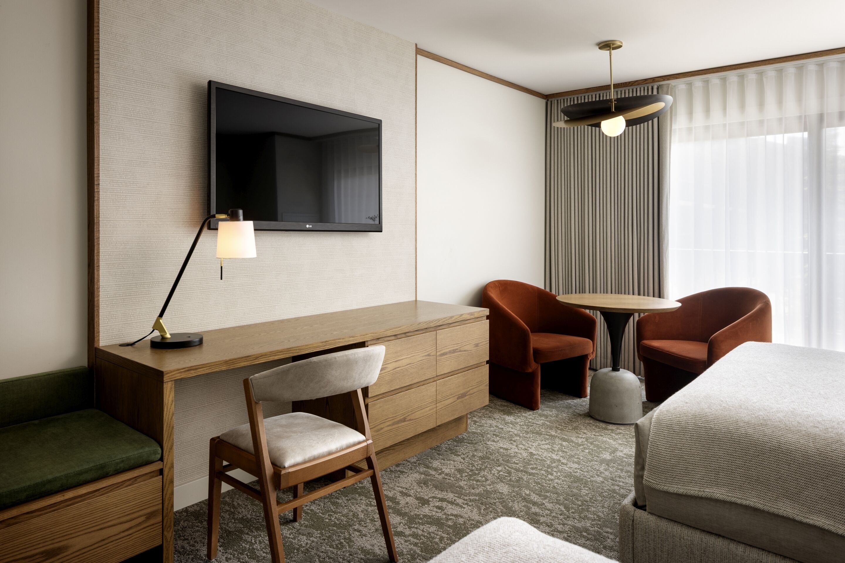 A hotel room with a wooden desk, a television mounted on the wall, two brown chairs, a round table, and a bed with a grey blanket.