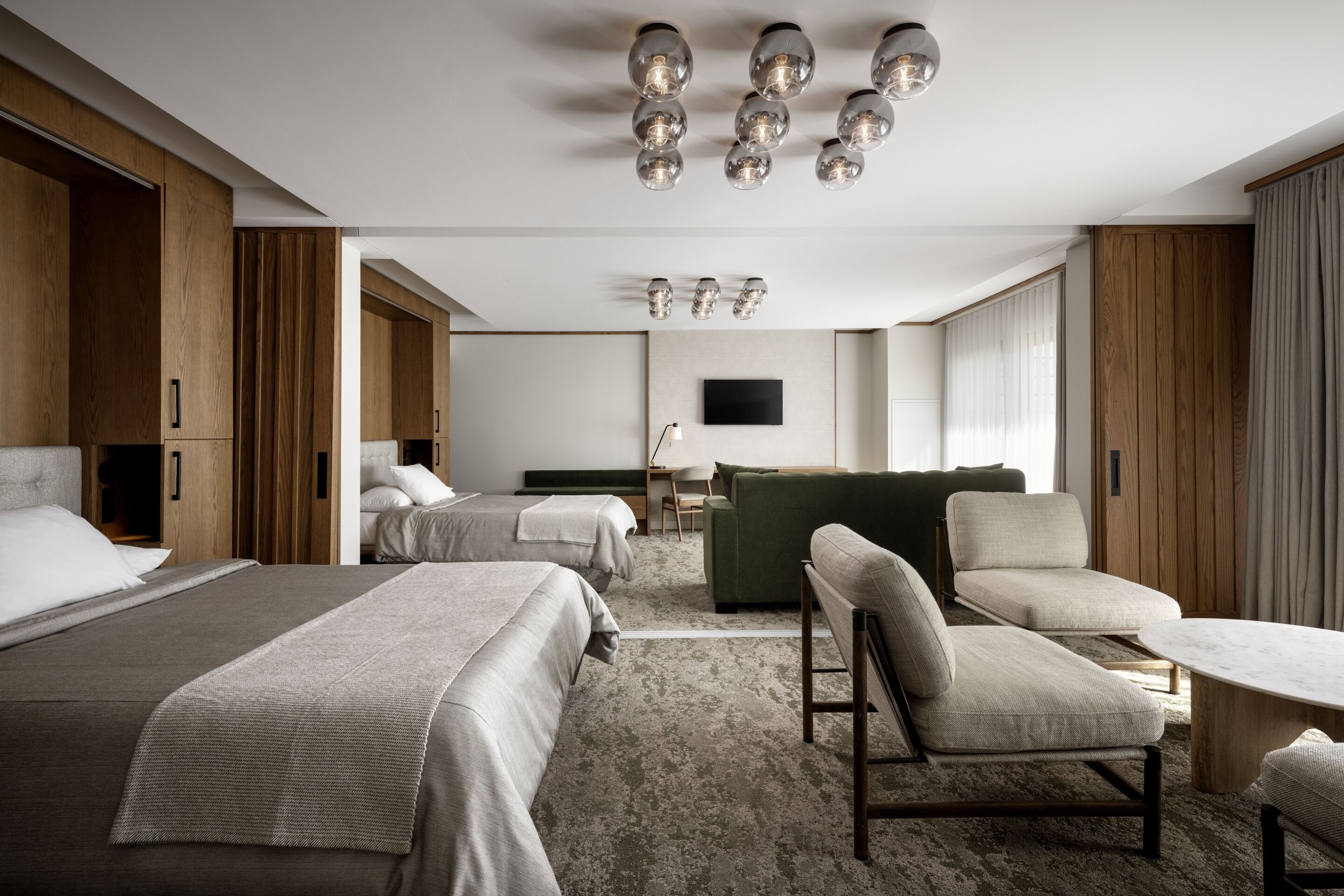 A modern hotel room with two beds, a green sofa, a small table, and a flat-screen TV mounted on the wall.