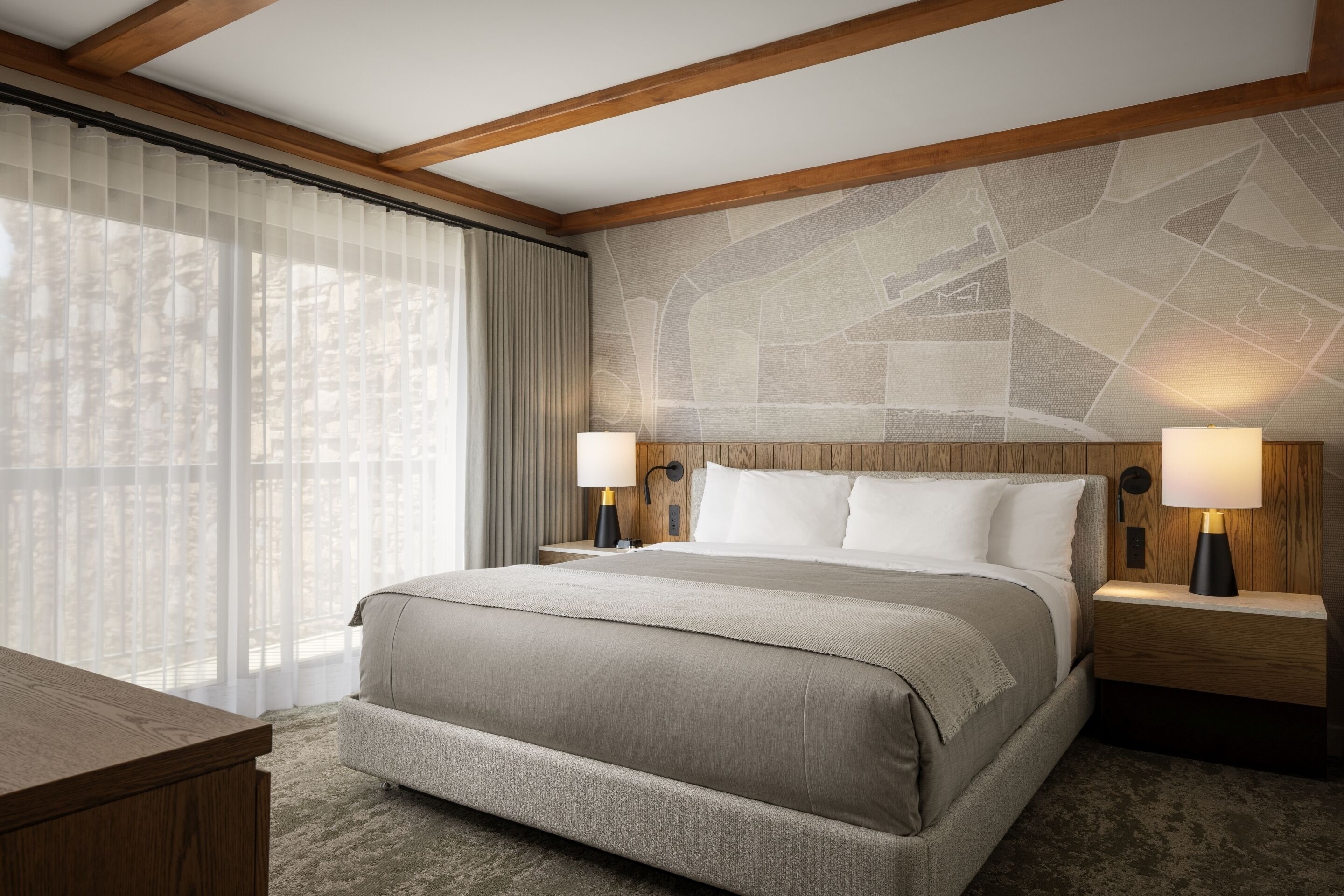 A hotel room with a large bed, wooden headboard, two bedside tables with lamps, a desk, and a view of trees through sheer curtains.