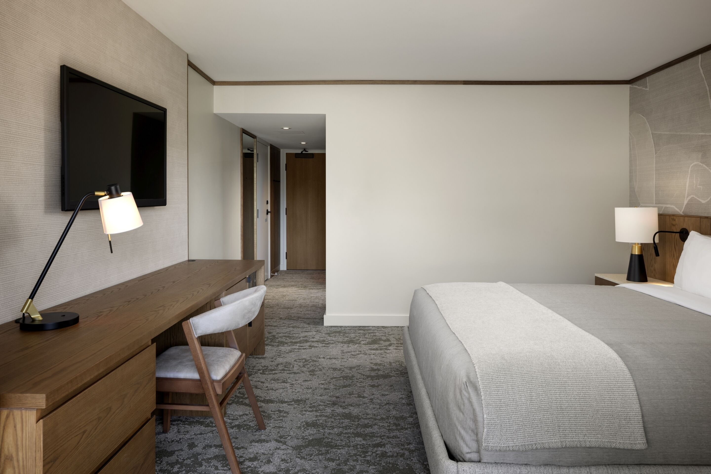 A hotel room with a wooden desk, a chair, a bed with a grey blanket, a TV mounted on the wall, and a lamp on the nightstand.