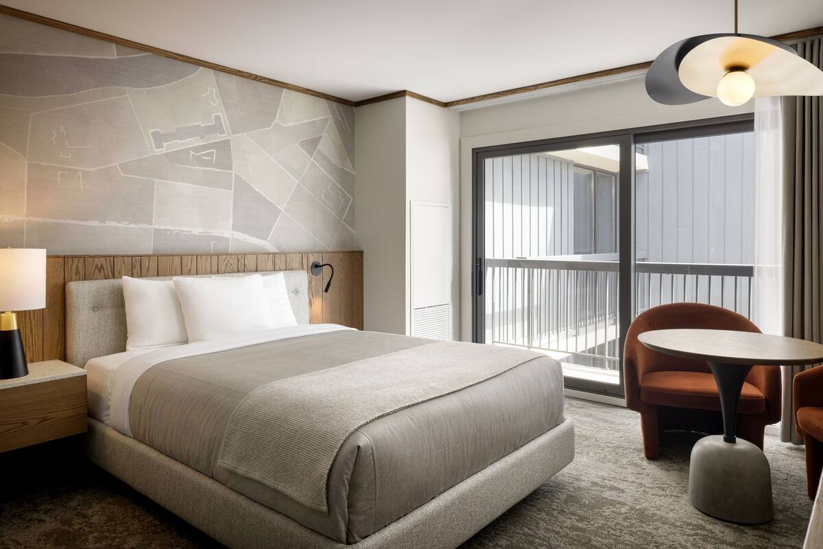 A hotel room with a large bed, a wooden headboard, a nightstand with a lamp, a round table with two chairs, and a view of the outside through a sliding door.