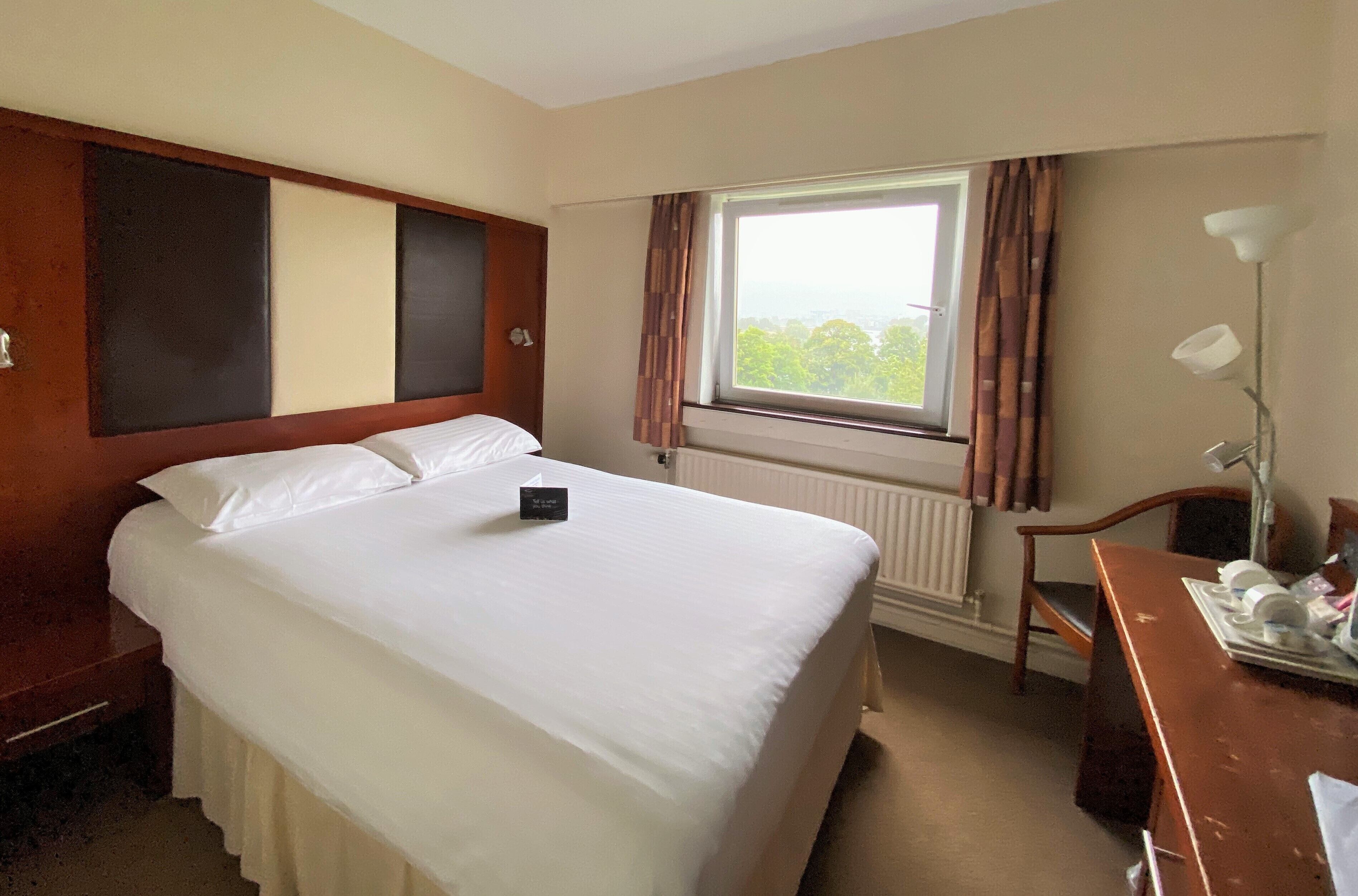 Une chambre d’hôtel avec un grand lit, un bureau, une chaise, une fenêtre avec des rideaux et un radiateur.