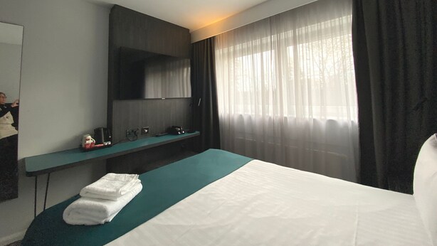 Une chambre d’hôtel avec un lit, un bureau, un miroir et une fenêtre avec des rideaux.