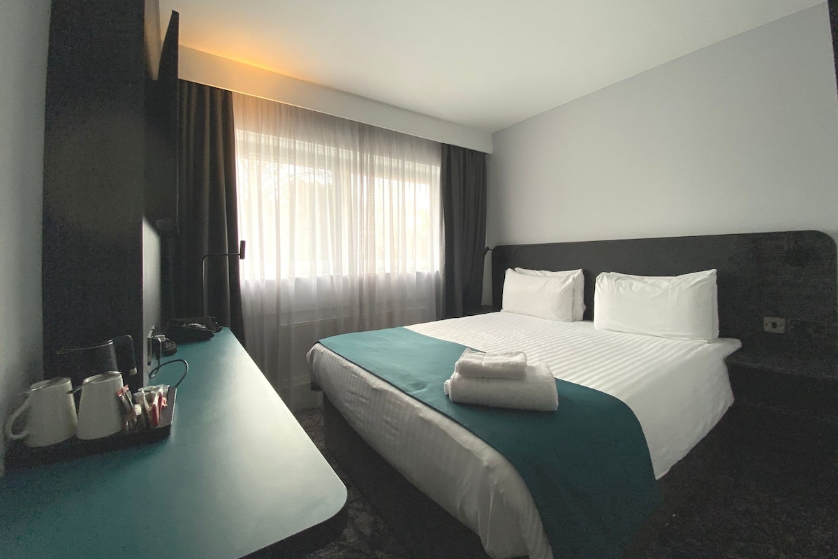 A hotel room with a bed, a desk with a chair, a coffee maker, and a window with curtains.